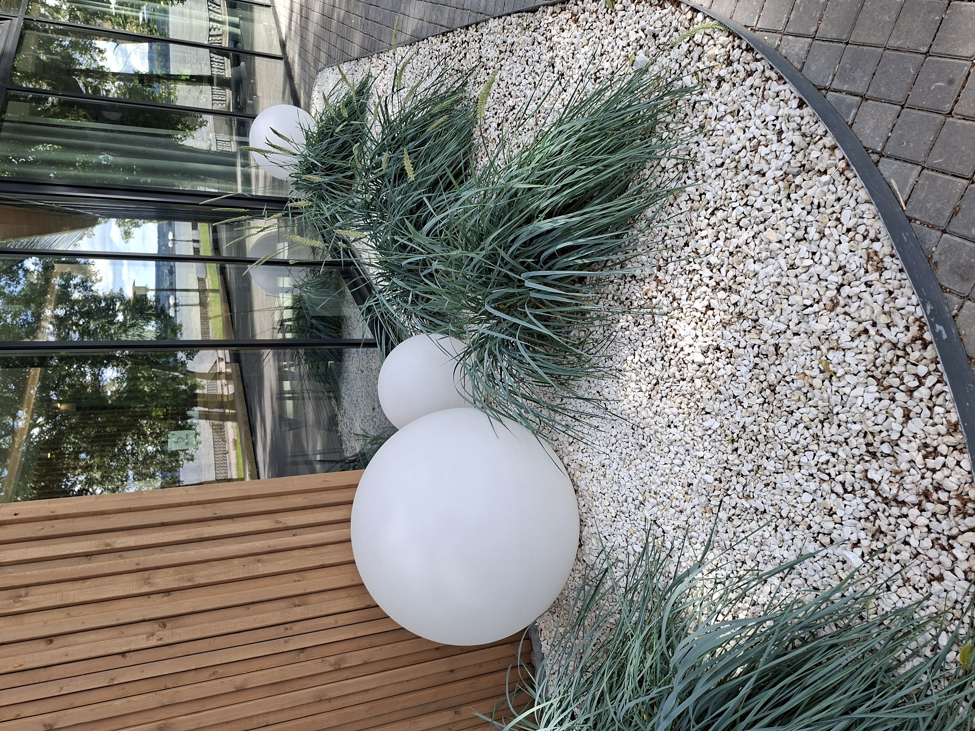 The image depicts an outdoor scene featuring a modern architectural element, a large spherical white light fixture, and ornamental grasses. The architectural element consists of a curved wall clad in vertical wooden slats, a floor paved with square gray tiles, and a large glass facade. The glass facade reflects trees, the sky, and a body of water. The spherical light fixture is positioned in a bed of small white gravel, alongside several clumps of blue-green ornamental grass with seed heads. A second, smaller spherical light fixture is partially visible behind a clump of grass. The setting suggests a contemporary public space, possibly a park or a plaza, adjacent to a body of water. The location is identified as being in the city of Votkinsk, Russia. There are no people visible in the image, and no actions or interactions are taking place.