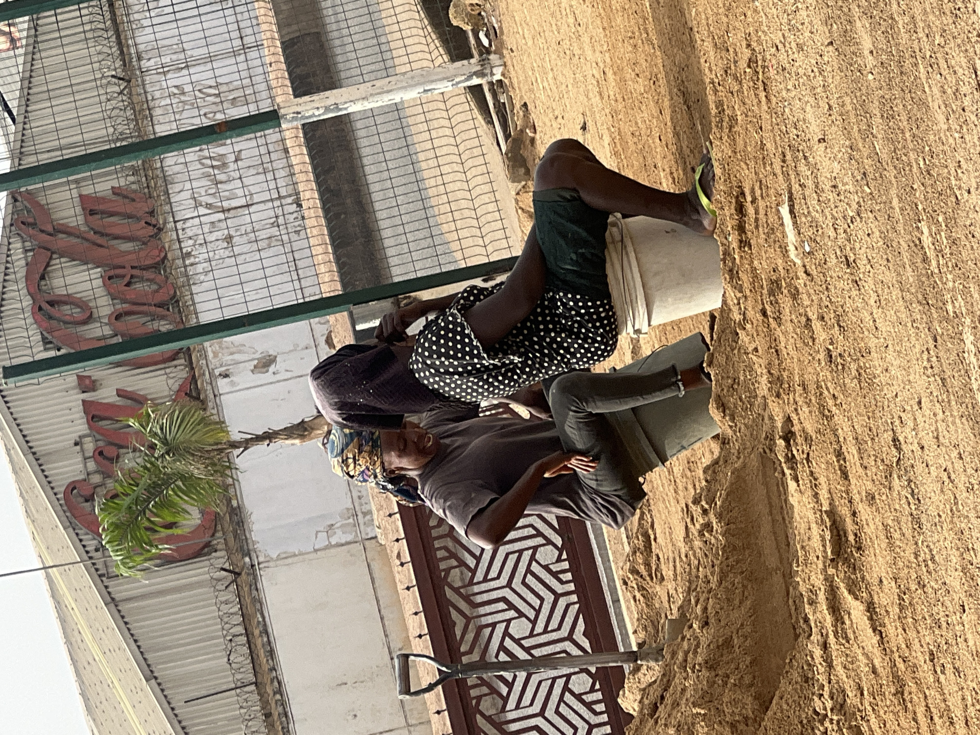 Two individuals are positioned near what appears to be a large pile of sand. One person is seated atop a white bucket, wearing shorts and a polka-dotted garment. The other is standing and wearing a dark shirt, head wrap, and jeans. A shovel rests near the sand pile and a decorative gate is behind the standing figure. The setting appears to be outdoors in proximity to a building. A sign is visible on the building. Based on the prompt, the location is Barakin Lerawi, Nigeria.