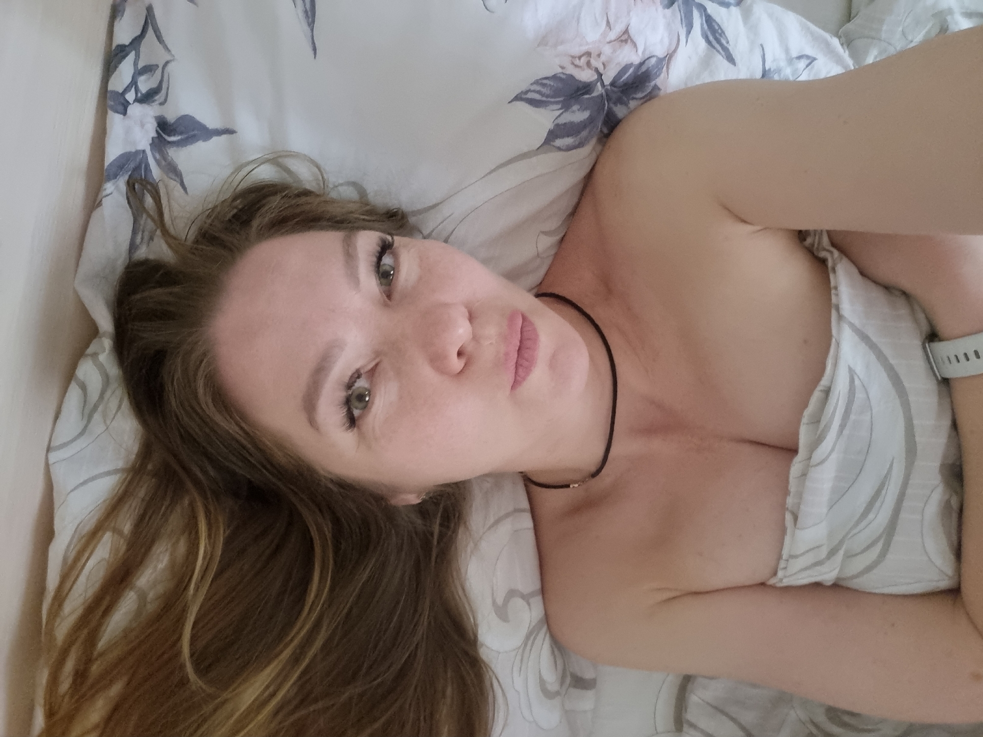 A woman lies in bed. Her head is turned to the right side of the image, and she looks towards the camera. She has long brown hair, fair skin, green eyes, and pink lips. She is wearing a thin black necklace. Her body is covered by a light-colored patterned bedsheet. The background consists of a pillow and wall. There are no other people or clear indicators that suggest the location is Chaikovsky, Russia.