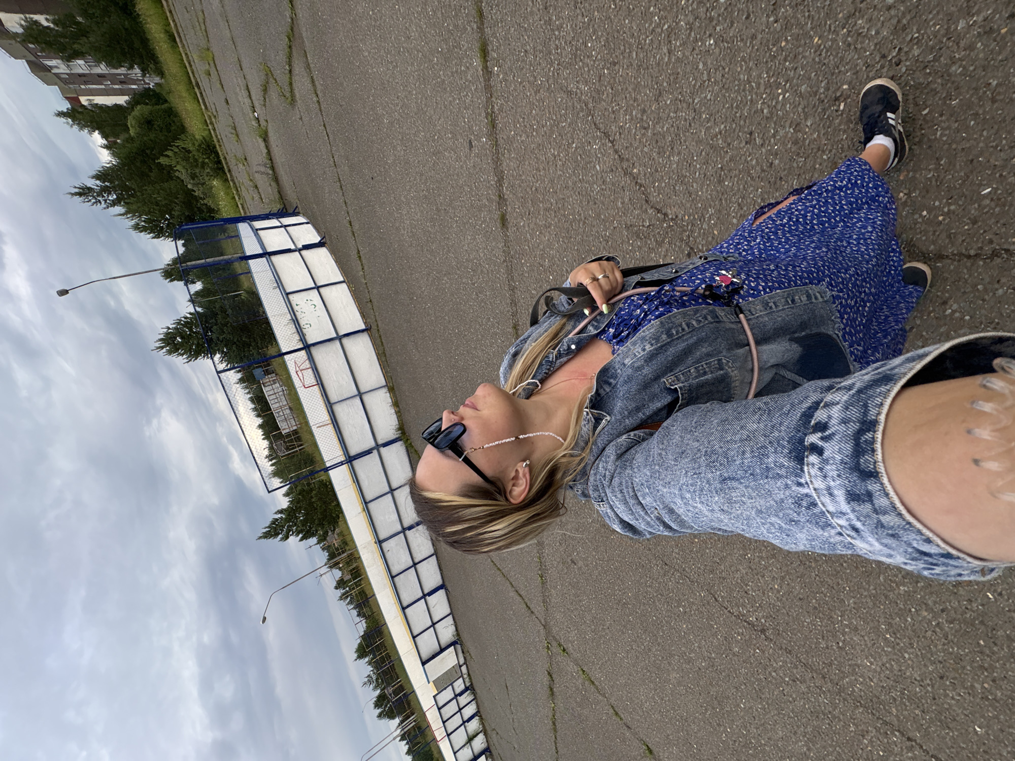 A woman is taking a selfie outdoors. She is wearing a blue floral dress, a denim jacket, sunglasses, and black sneakers with white socks. She has a necklace made of small white beads and a ring on her left hand. Her toenails are painted green. The scene is an outdoor paved area next to a fenced sports court, possibly a hockey rink, with a basketball hoop visible at one end. Trees and a building can be seen in the background. The sky is overcast, suggesting a cloudy day. The woman's expression is neutral, and the overall mood is casual. The location is identified as Марковский, Russia. There is no visible text in the image.