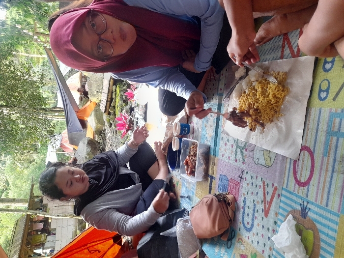 Three people sit on a patterned mat outdoors, seemingly camping. One person wears a maroon hijab and glasses, another a black scarf. Food, including noodles and skewers, is laid out on paper. A clear plastic container of food sits nearby, along with drinks and a brown handbag. Tents are visible in the background, surrounded by trees. The focus is on the group enjoying their meal in a casual, outdoor setting.