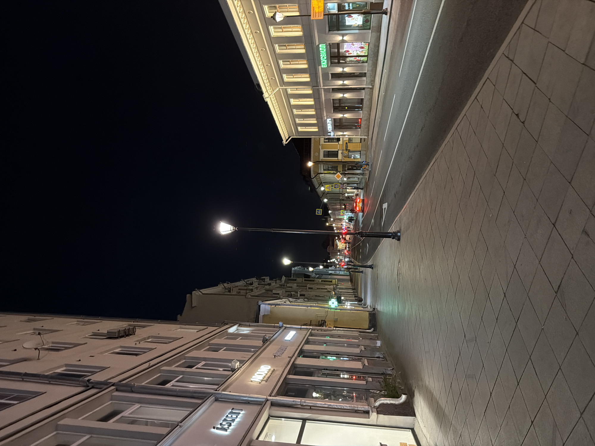 A nighttime view of a pedestrian sidewalk and street in Moscow, Russia. The sidewalk is paved with square tiles and is lined with buildings on the left. The buildings appear to be shops or offices, with illuminated signs visible. The street is paved with asphalt and has white lane markings. Streetlights illuminate the scene. A few cars are visible on the street, and one red car is moving. The sky is dark, indicating it is night. The overall impression is of a quiet, urban street scene at night.