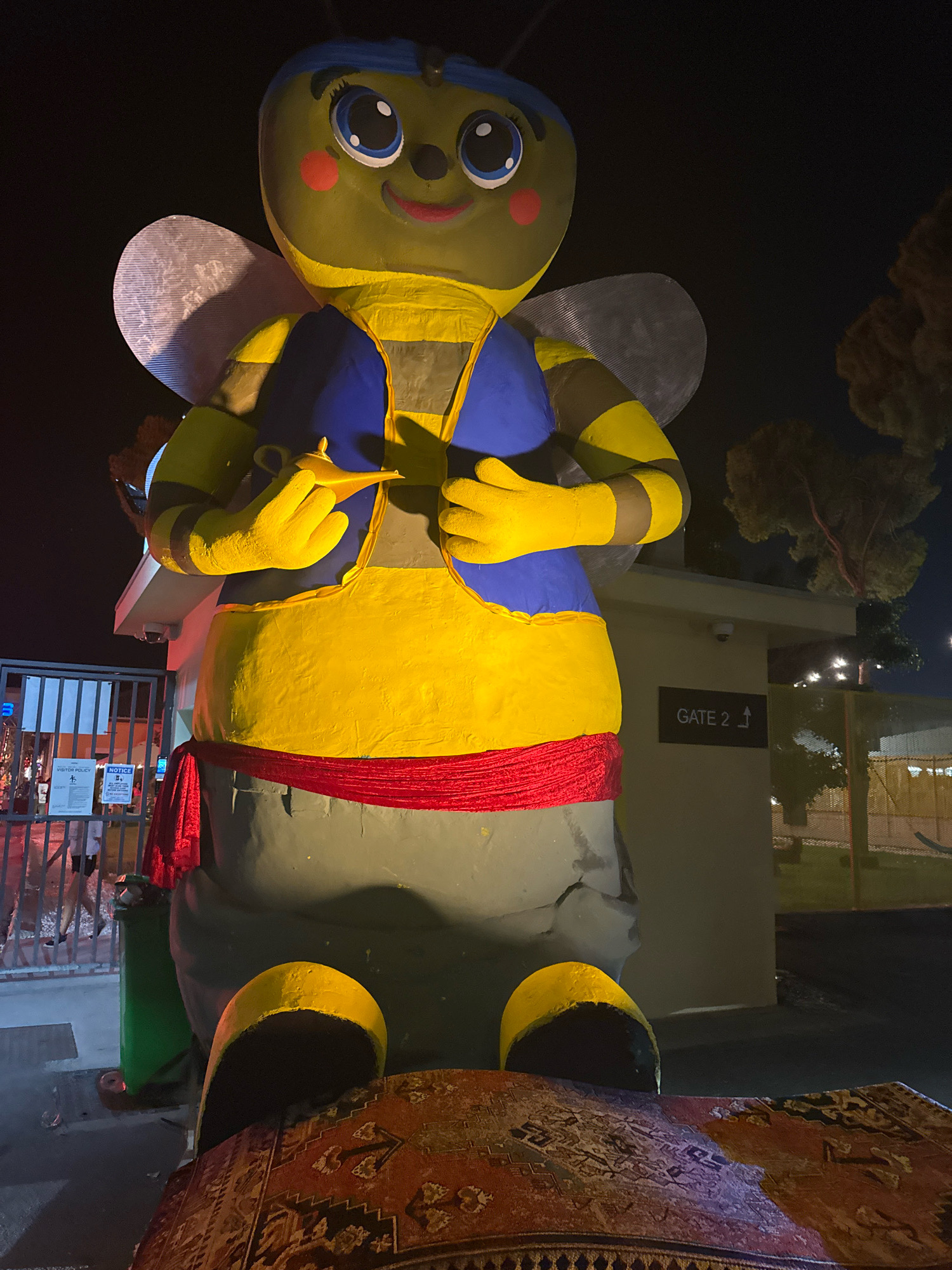 A large, yellow, cartoonish bee mascot is standing in what appears to be a nighttime outdoor setting, possibly an event entrance or theme park. The bee has large blue eyes with black pupils, red rosy cheeks, and a blue headband. It is wearing a blue vest over a yellow and black striped torso, and a red sash around its waist. The bee is holding a golden magic lamp in its hands. In the foreground, a portion of an ornate rug is visible. Behind the bee, a building with a sign that reads "GATE 2" is visible, along with a metal fence and some trees. The lighting suggests artificial illumination, and the background is dark, indicating nighttime. There are no other discernible people or interactions in the immediate vicinity of the mascot. The presence of a "GATE 2" sign might suggest a venue or a specific section within a larger area. The overall scene is festive and whimsical, typical of a mascot at an amusement or entertainment venue. It is not possible to determine the specific location in Limassol, Cyprus from the provided image.