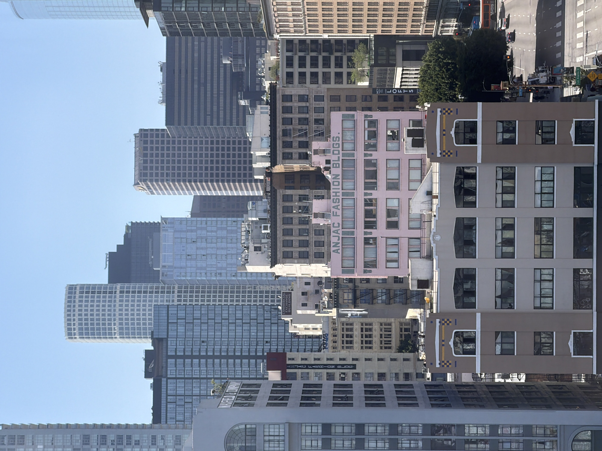 The image shows a cityscape with numerous buildings of varying heights and architectural styles. The buildings are densely packed, creating a layered effect, against a clear blue sky. A mix of old and new structures is visible, with some buildings having modern glass facades, while others have more traditional brick or stone exteriors. One building in the middle ground is painted a distinctive pink color with signage reading "ANJAC FASHION BLDGS." In the upper right, there is a view of a street with traffic, including cars and possibly buses. Based on the architectural style and building density, it is likely a central area of Los Angeles, United States of America.