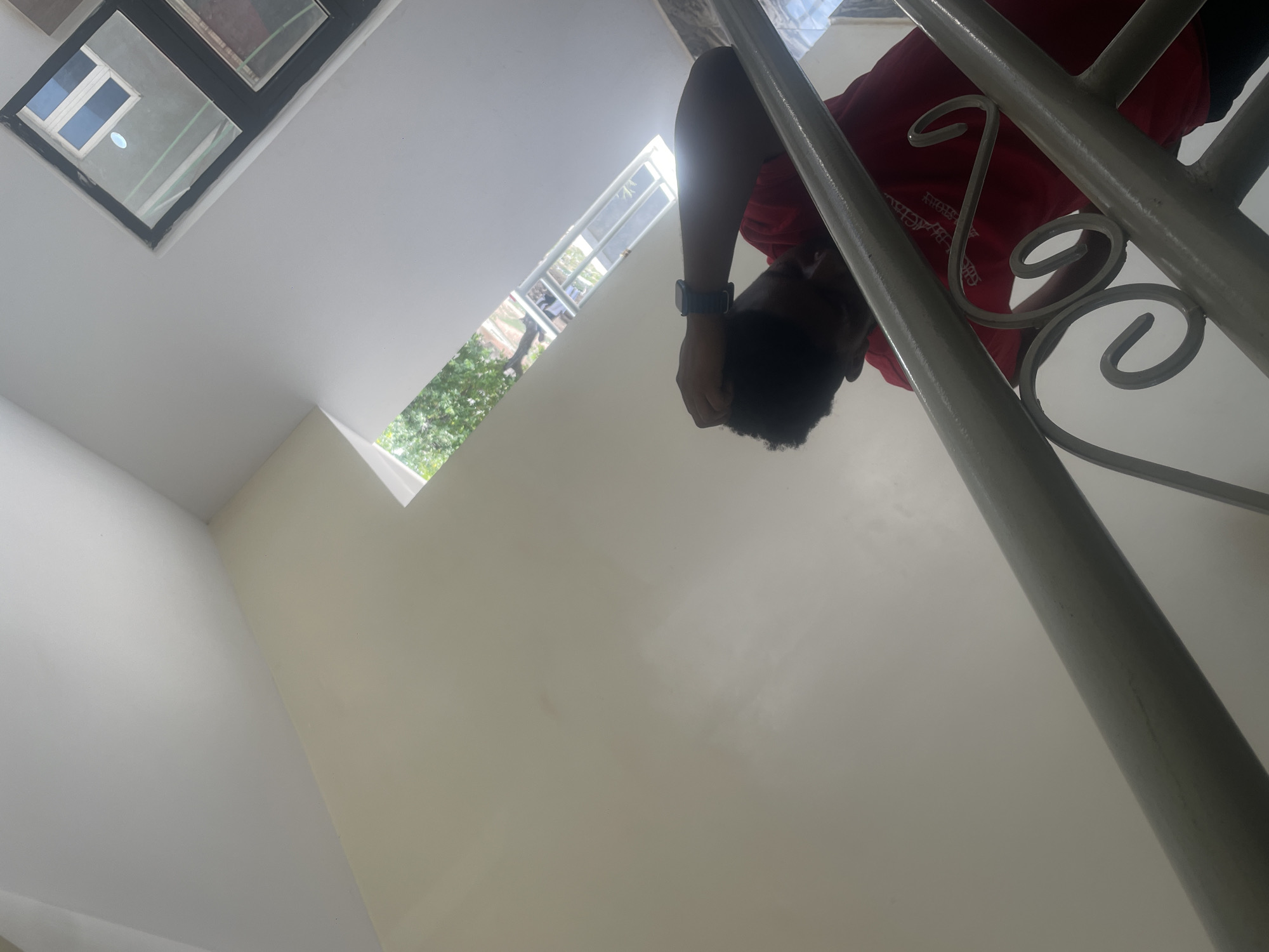 This upward-angled photograph, taken in Gwarinpa, Nigeria, depicts an interior view of a building, likely a stairwell or landing. At the top of the frame, a person, identified as male, leans over a light-colored, decorative stair railing. He is wearing a red short-sleeved t-shirt and a dark smartwatch on his left wrist. His head, with dark, short hair, is visible, and he appears to be looking down towards the camera.
The scene is dominated by clean, light-colored walls, appearing off-white or beige, and the elegant railing. The railing features several decorative, S-shaped scroll elements, painted in a light gray or white. Natural light floods the space from two windows. One rectangular window is situated high up on a wall, while another long, horizontal window provides a glimpse of vibrant green foliage and trees outside, confirming a bright daytime setting.
The perspective of the shot suggests the viewer is standing at a lower level, looking up at the person and the architectural details. The building appears modern and well-maintained. Partially legible white text can be seen on the red t-shirt, possibly part of a logo or design. The overall impression is one of brightness and spaciousness, illuminated by natural daylight.