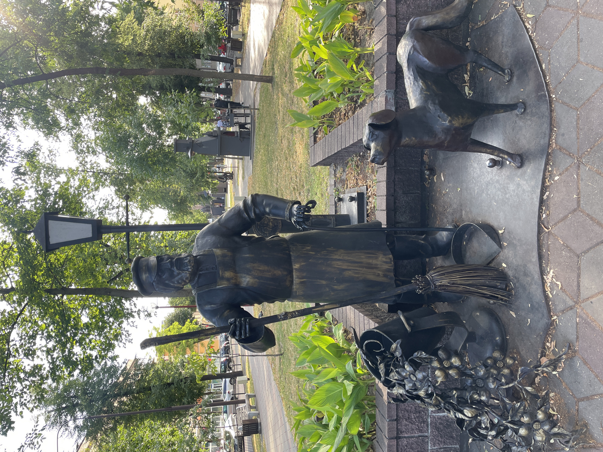 The media file shows a bronze sculpture of a street sweeper and a dog. The street sweeper is depicted in a uniform, holding a broom and dustpan. The dog is standing beside the sweeper. The sculpture is placed in an outdoor setting with trees and greenery visible in the background. The ground around the sculpture is paved with bricks.
The scene appears to be a park or a public space, possibly in a city, given the presence of trees, benches, and a sidewalk. The lighting suggests it is daytime, with sunlight casting shadows. The weather appears to be pleasant, suitable for outdoor activities.
Notable details include the intricate details of the sculpture, such as the clothing of the street sweeper, the fur of the dog, and the leaves of the plants. The sculpture itself is the primary subject, with the background providing context to its location. The overall mood conveyed by the sculpture is one of diligence and companionship.
There is no visible text in the media file. The location context is Brest, Belarus.