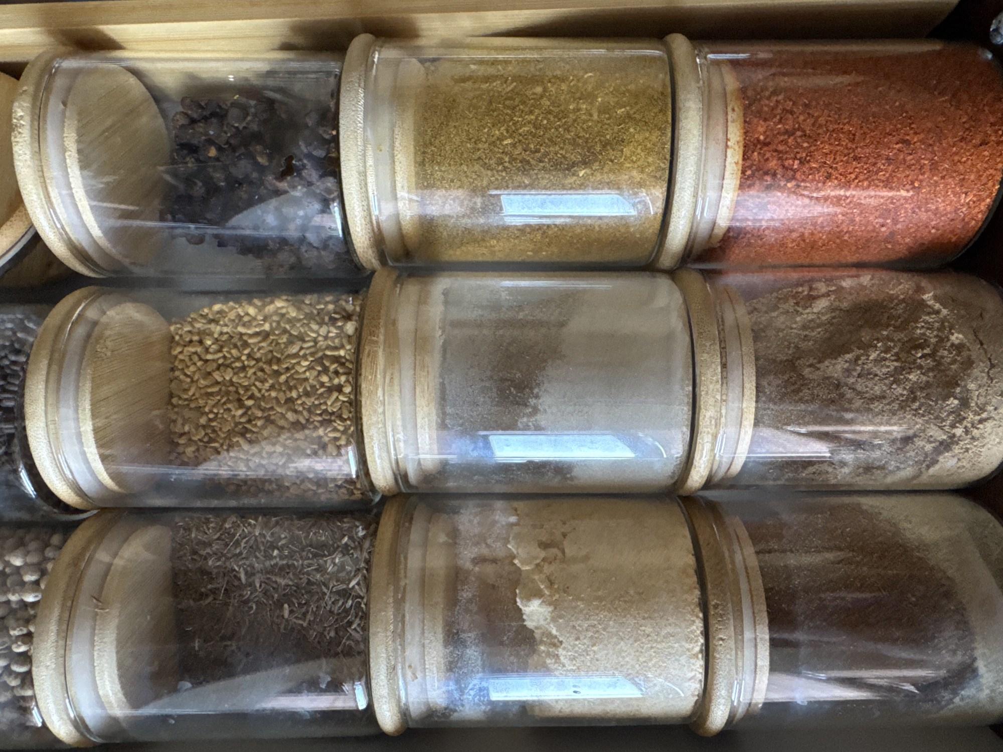 A collection of clear glass spice jars with bamboo lids are neatly arranged in a grid on a wooden shelf. The jars contain various spices. In the top row, from left to right, are whole cumin seeds, fenugreek seeds, and whole black peppercorns. The middle row, from left to right, contains a pale yellow powder, a white powder, and a golden yellow spice blend. The bottom row, from left to right, shows a dark brown powder, a light brown powder, and red chili flakes. The scene appears to be indoors, likely in a kitchen or pantry. The lighting is bright, and the jars reflect the light.