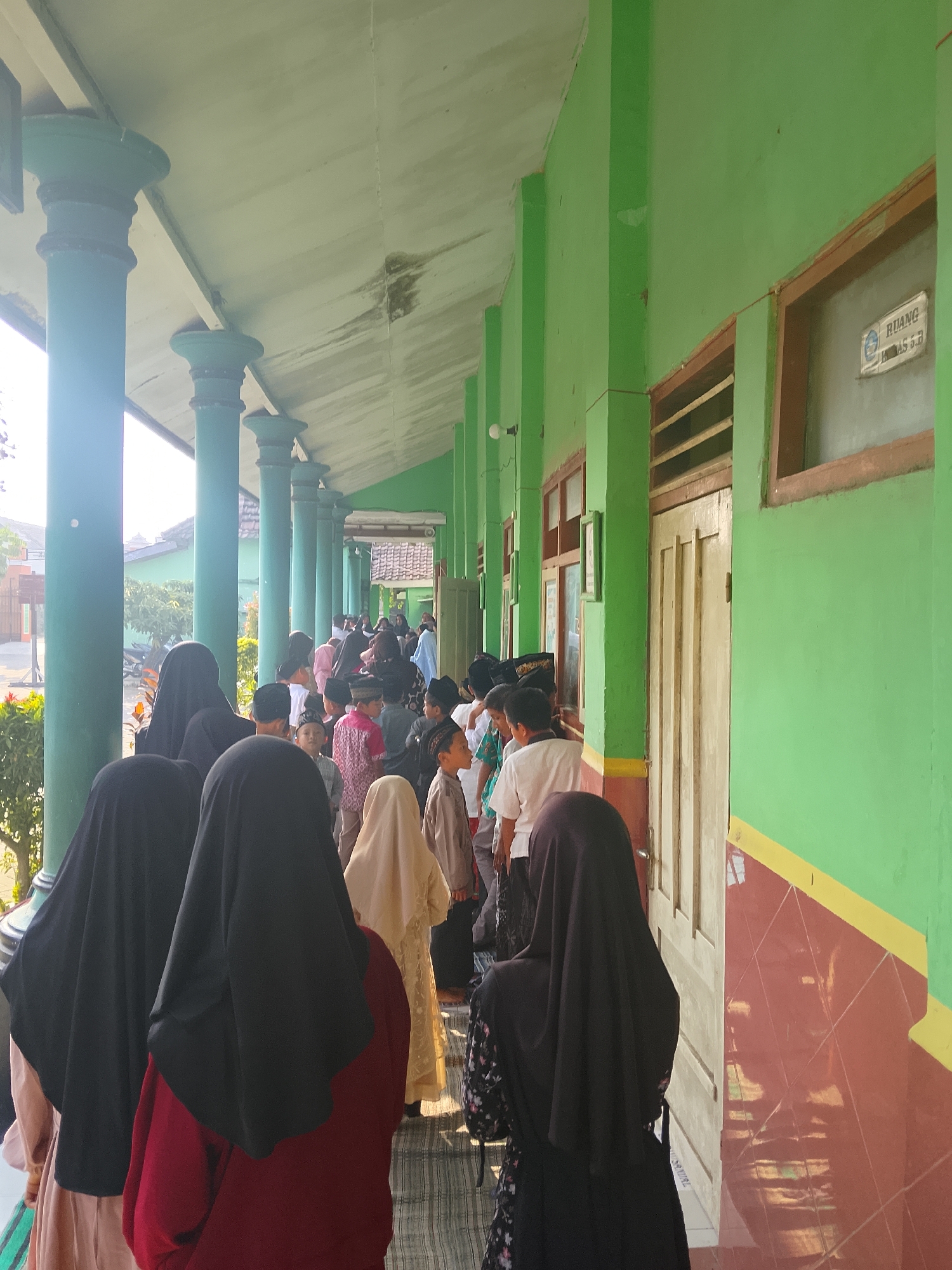 The image depicts a covered walkway outside a building with green walls and pillars. A group of children and adults, many wearing hijabs or peci caps, are standing in a line. The building appears to be a school or religious institution, with signs on the doors indicating "RUANG KELAS 6.B" (Classroom 6.B). Sunlight streams in from the left, casting shadows. The ground is tiled. The overall atmosphere is one of an organized gathering or activity. Based on visual cues, this scene is likely located in Kepuh, Indonesia, given the architectural style and attire of the people.