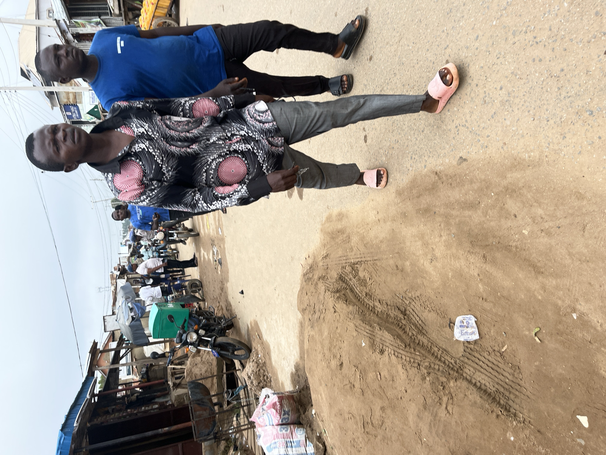 Two men walk on a dusty road in what appears to be an informal market area in Abuja, Nigeria. The man in the foreground, on the left, wears a patterned short-sleeved shirt with black, white, and pink designs, grey trousers, and pink slip-on sandals. He is looking slightly to his left. To his right and slightly behind him, a second man in a blue t-shirt and black trousers walks, looking forward. In the background, various market stalls with corrugated metal roofs are visible, along with motorcycles, bags of what appears to be cement, and piles of sand. The ground is uneven and appears to be unpaved. The sky is overcast.