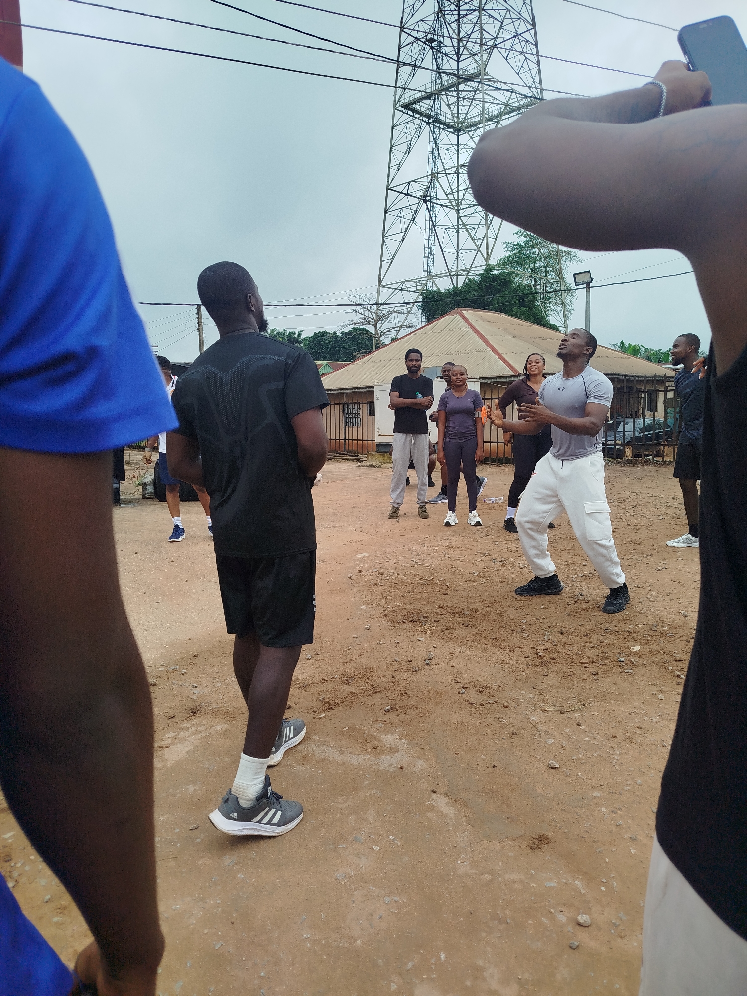 In Uselu, Nigeria, a group of people are gathered outdoors on a dirt ground. A man in a black t-shirt and black shorts stands facing away from the camera, with his left arm slightly raised. He is wearing white socks and grey and white athletic shoes. To his right, a man in a grey t-shirt and white pants is facing the camera, with his arms extended in front of him and his eyes closed. He appears to be leading an exercise or activity, with a few other individuals watching him. In the background, a large metal tower structure is visible against a cloudy sky, along with some buildings and trees. The scene suggests an outdoor fitness or group activity.