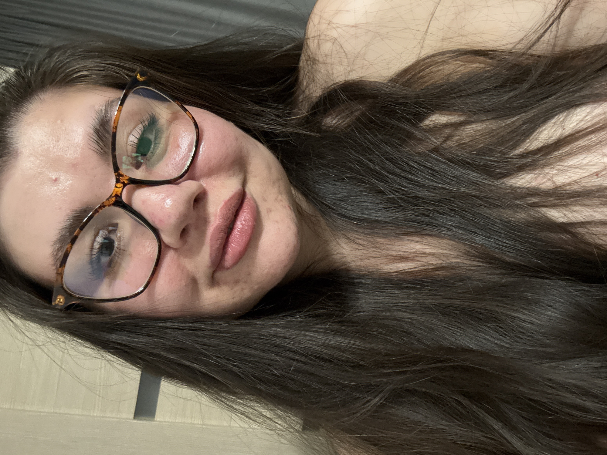 A woman with long brown hair and glasses is lying down. Her right eye is reflected in the glasses. The background is a light-colored wall. There is no indication that this image was taken in Votkinsk, Russia.