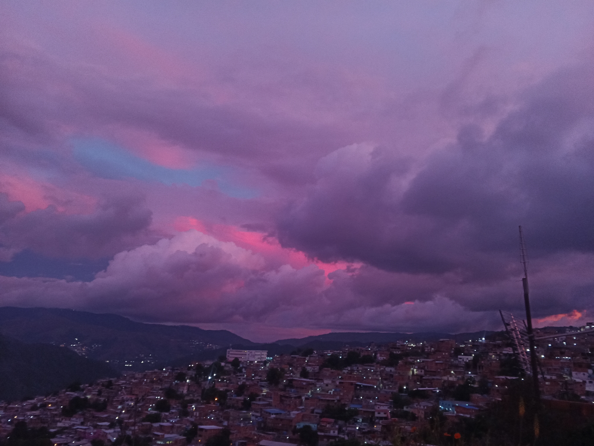 This is a horizontal, medium-range shot of a densely populated hillside neighborhood in Caracas, Venezuela, at dusk. The sky is a dramatic display of purple and pink clouds, with some areas of light blue peeking through. The city lights are beginning to twinkle across the hillside, illuminating the rooftops of numerous brick and concrete buildings. In the background, dark, rolling hills are visible, fading into the twilight. A tall, thin metal tower, possibly a cell tower or antenna, stands prominently on the right side of the frame. The overall atmosphere is moody and colorful, characteristic of a tropical sunset.
