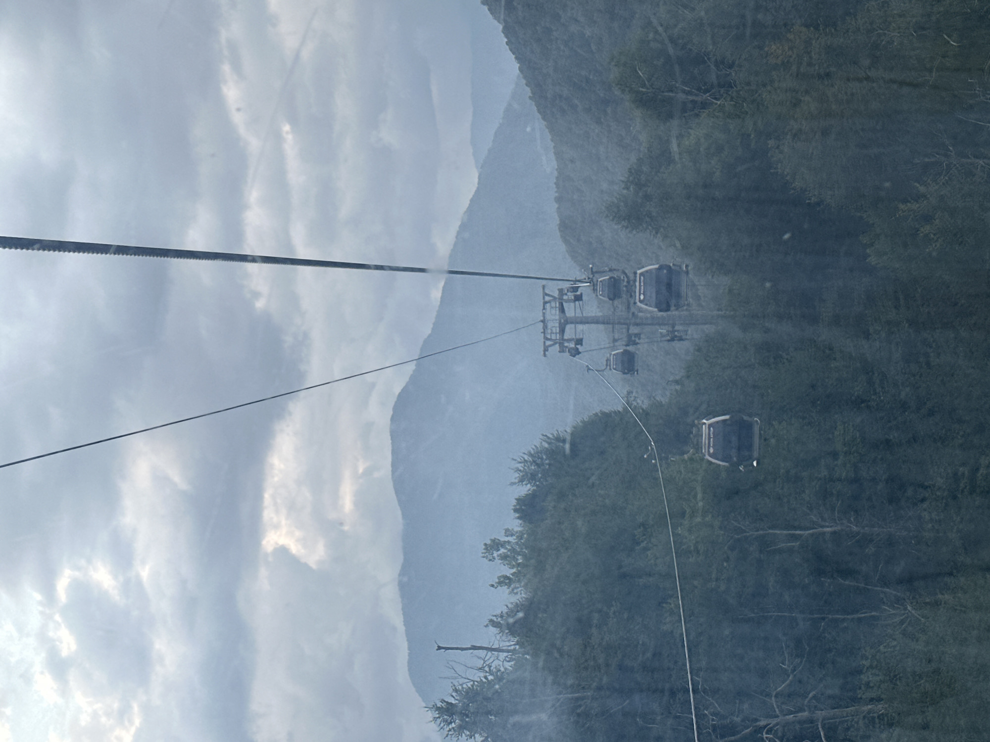 An aerial tramway operates against a backdrop of misty mountains and dense, dark green forest. The tramway consists of a cable and multiple gondolas, some of which are visible and suspended from the cable. The gondolas are modern and appear to be enclosed. The sky is overcast with a mix of dark and lighter gray clouds, suggesting a potentially moody or rainy atmosphere. The mountains in the background are layered and appear to recede into the distance, with the mist softening their outlines. The foreground is dominated by the lush, dark foliage of trees, creating a sense of depth and immersion within the natural landscape. The scene captures a moment of transit within a mountainous, possibly forested region in Estosadok, Russia.