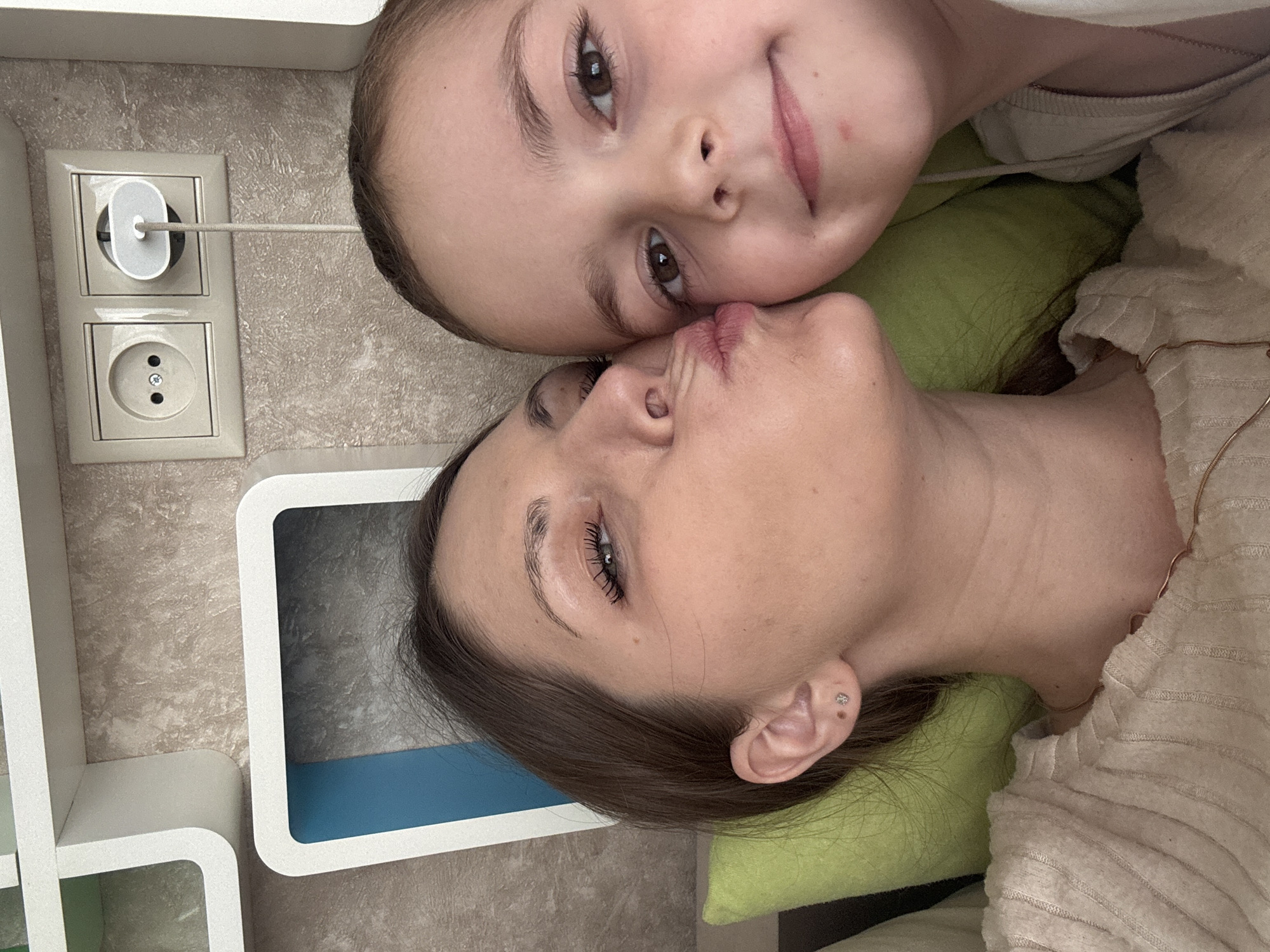 A woman and child lie head-to-head on a light green pillow. The woman is older, with fair skin, brown hair pulled back, prominent eyelashes, and a silver stud earring in her left ear. Her lips are pursed in a kissing gesture. She wears a light-colored ribbed top. The child is younger, with fair skin and brown hair. The child smiles slightly.
The background features a wall with light tan textured wallpaper and a European-style outlet with a white charging plug. To the left, there's a white shelving unit with a blue interior in one compartment.
It is impossible to confirm the location as Раздолье, Russia.