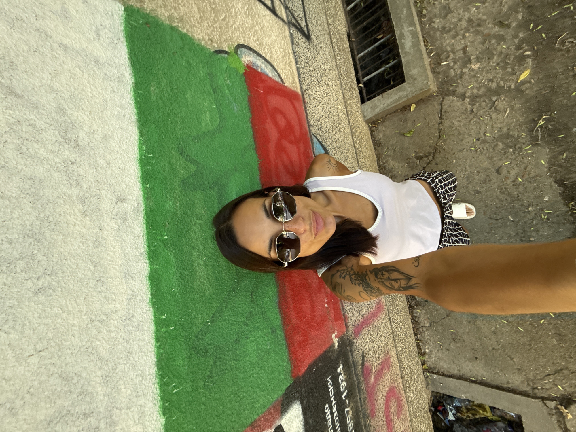 In the city of Pleven, Bulgaria, a woman takes a selfie in front of a wall painted with the Bulgarian flag. The wall is textured and appears to be made of concrete. The flag's colors, white, green, and red, are visible in horizontal stripes. The woman is wearing sunglasses, a white tank top, and patterned shorts. Her left arm is visible, displaying several tattoos. To the left of the woman, the wall has some faded text and graffiti. The ground in front of the wall is paved with concrete and shows signs of wear, with some scattered leaves. To the right, a metal grate covers a drainage channel. The scene appears to be outdoors, likely in an urban environment.