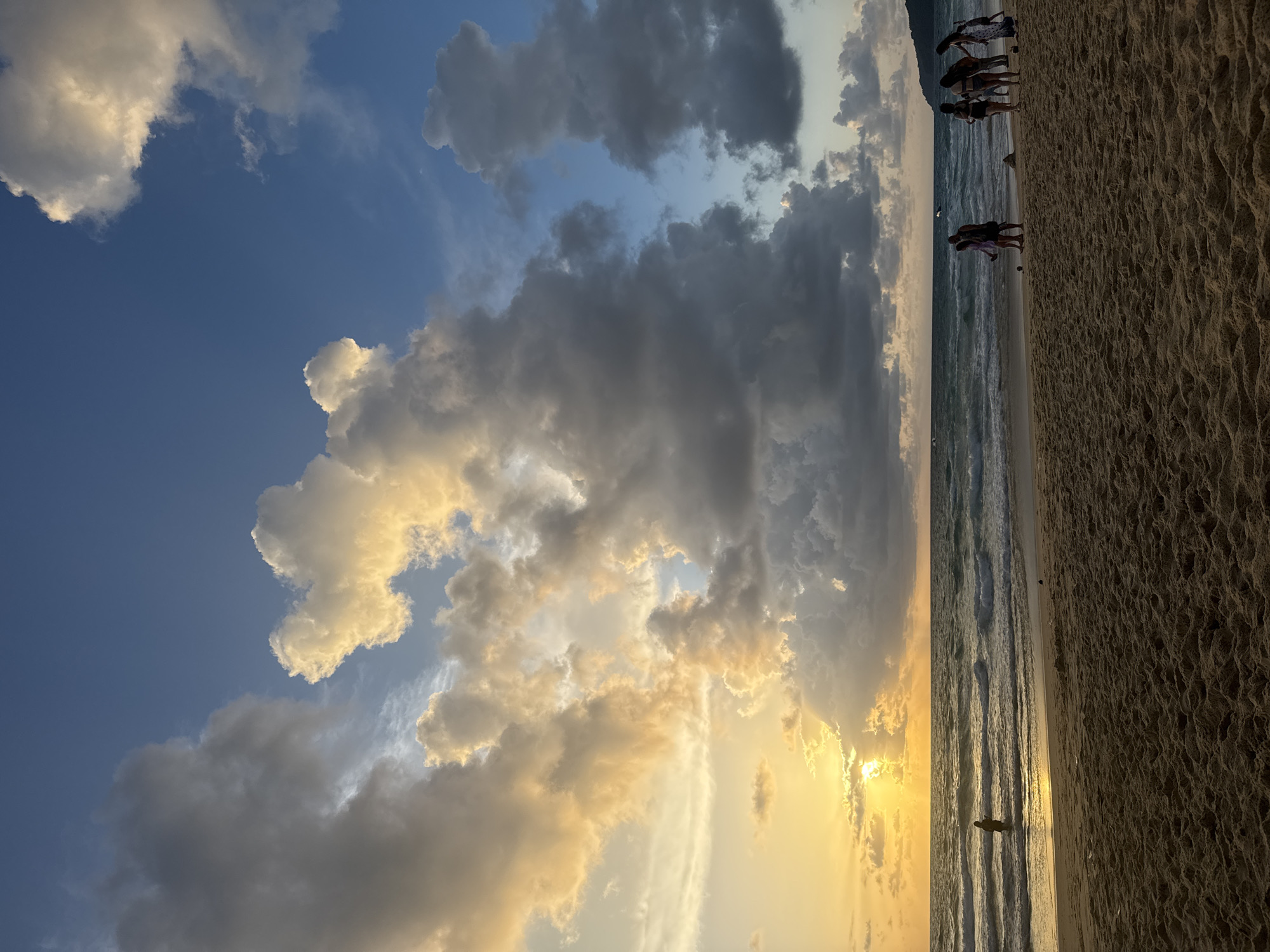 A wide shot captures a beach scene in Karon, Thailand, during a sunset. The sky is a dramatic canvas of blues and grays, with large, illuminated clouds catching the golden light of the setting sun. The sun, partially obscured by clouds, casts a warm glow on the horizon and the ocean's surface, creating shimmering reflections. Gentle waves roll onto a sandy beach, where a few individuals are visible. In the foreground, the sand is textured with subtle ripples and patterns. Further down the beach, a group of people are gathered, some standing and observing the sunset, while others appear to be engaged in an activity. One person is wading in the shallow water near the left side of the frame. The overall atmosphere is serene and picturesque, characteristic of a tropical beach sunset.