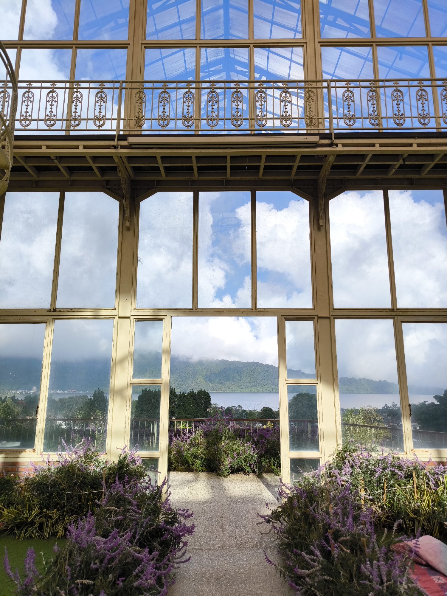 The image shows an interior view of a building in Candikuning, Indonesia, featuring large windows that offer a view of a lake surrounded by mountains. The building has a balcony with an ornate metal railing. Below, the interior space opens to the outside, with the view framed by the building's structure. Purple flowers and other greenery are planted in front of the building. The windows reflect the sky and clouds, creating a sense of depth.