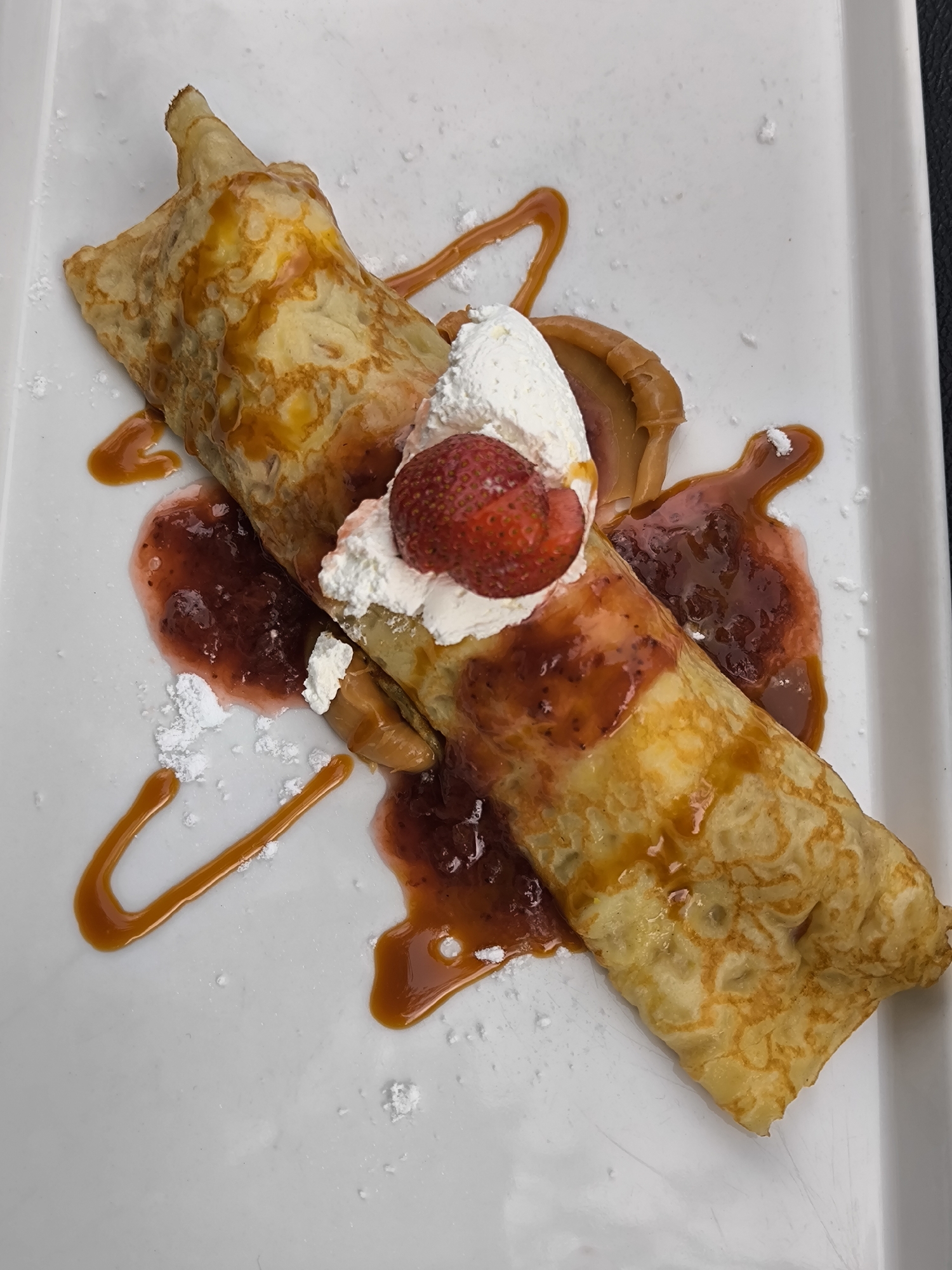 On a white rectangular plate sits a crêpe dessert. The crêpe is rolled and topped with whipped cream and a sliced strawberry. Strawberry jam and caramel sauce are drizzled around the crêpe. Powdered sugar is sprinkled on the plate. The image was taken in Mekarsari, Indonesia.