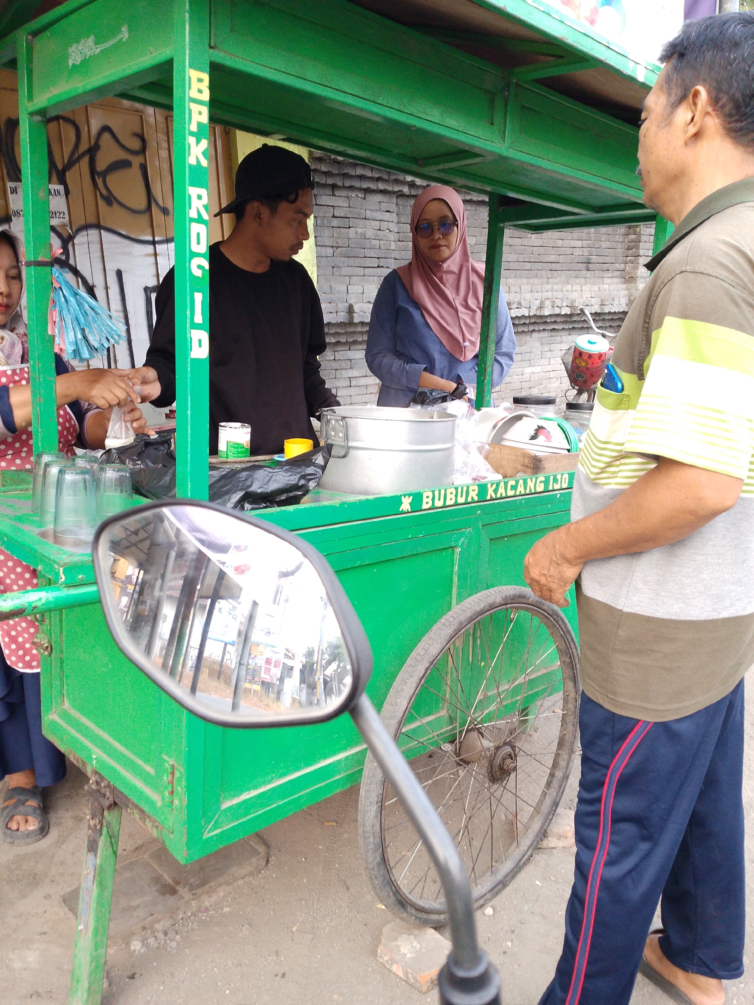 In Tegalwangi, Indonesia, two individuals, a man wearing a black cap and a woman in a pink hijab, stand behind a bright green food cart. The cart is adorned with the text "BPK RKSID" vertically on one of its support beams and "BUBUR KACANG IJO" horizontally across the front. The man appears to be preparing a drink, holding a plastic bag that contains a white liquid. The woman is in the process of handing over something, possibly money or an item, to the man. In the foreground, a motorcycle's rearview mirror reflects the scene, showing a street with buildings and a clear sky. A man in a striped t-shirt and blue track pants stands to the right of the cart, facing the two individuals. The cart itself is on wheels, suggesting it is mobile. The background reveals a brick wall and some graffiti.