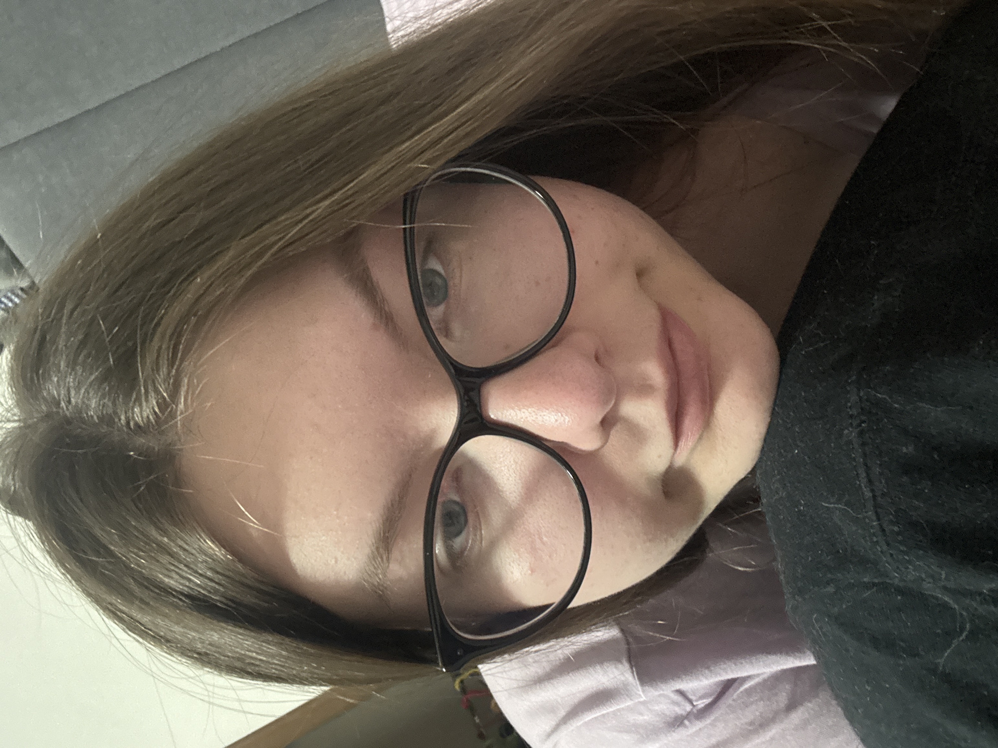 A young woman with light brown hair and glasses is looking down at the camera. She is wearing a light purple t-shirt and a black jacket. The background is blurry and appears to be a room with a grey wall. There is no information about the location being Kyiv, Ukraine.