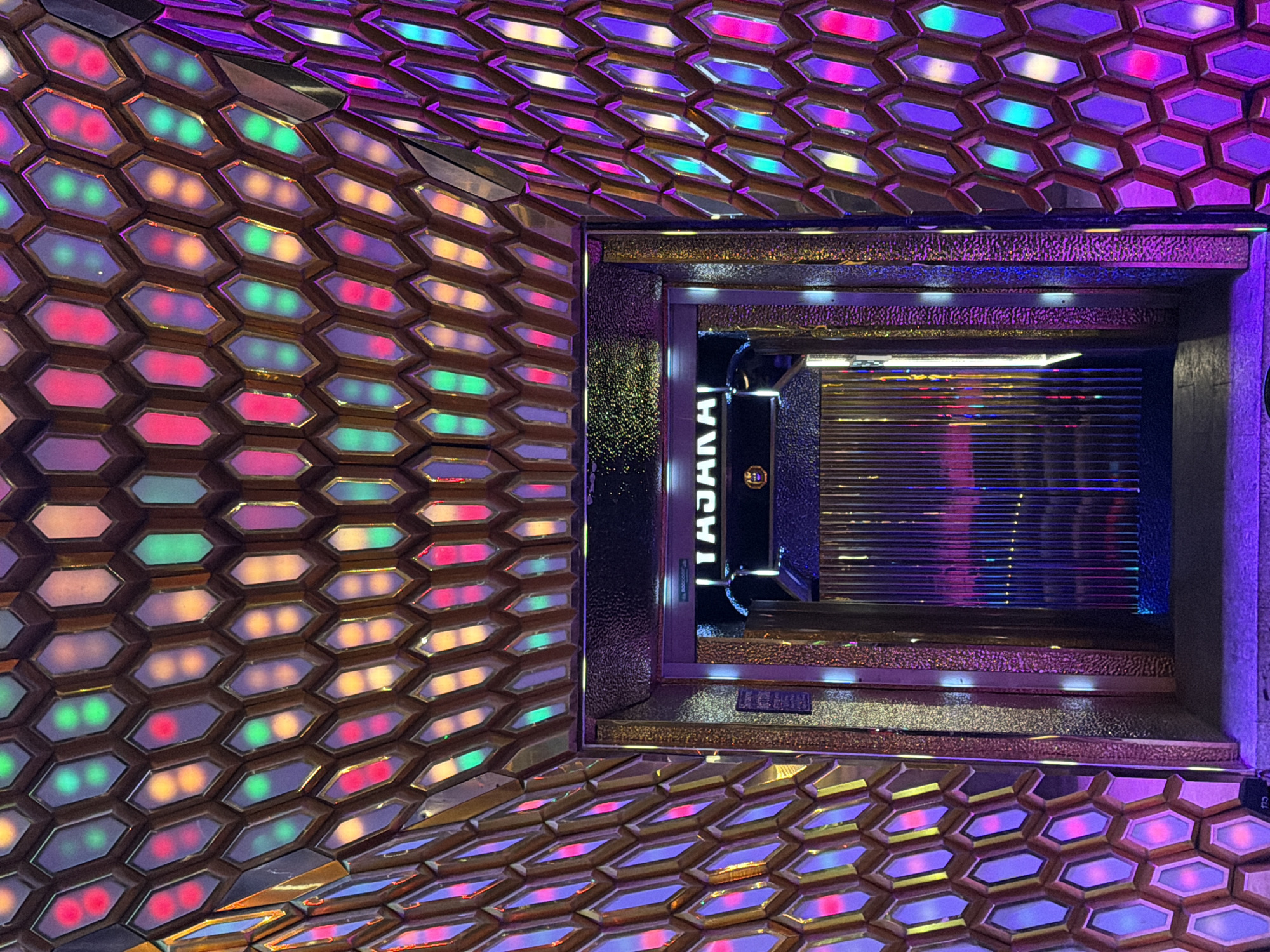 The image captures the entrance to a venue adorned with a honeycomb-like pattern of hexagonal panels, each illuminated with vibrant, multi-colored lights. The overall aesthetic is futuristic and dynamic, with a strong emphasis on lighting effects. The entrance itself is framed by ornate, gold-toned metallic elements and textured surfaces, leading into a dimly lit interior featuring a set of vertical, reflective panels that suggest a stage or performance area. Above the entrance, a sign reads "TAJMAHAL" in bold, capital letters, suggesting the name of the establishment. The floor in front of the entrance is paved with dark material, and the surrounding walls and ceiling continue the hexagonal illuminated panel motif, creating an immersive visual experience. The scene is set in Nha Trang, Vietnam, a coastal city known for its vibrant nightlife and entertainment venues.