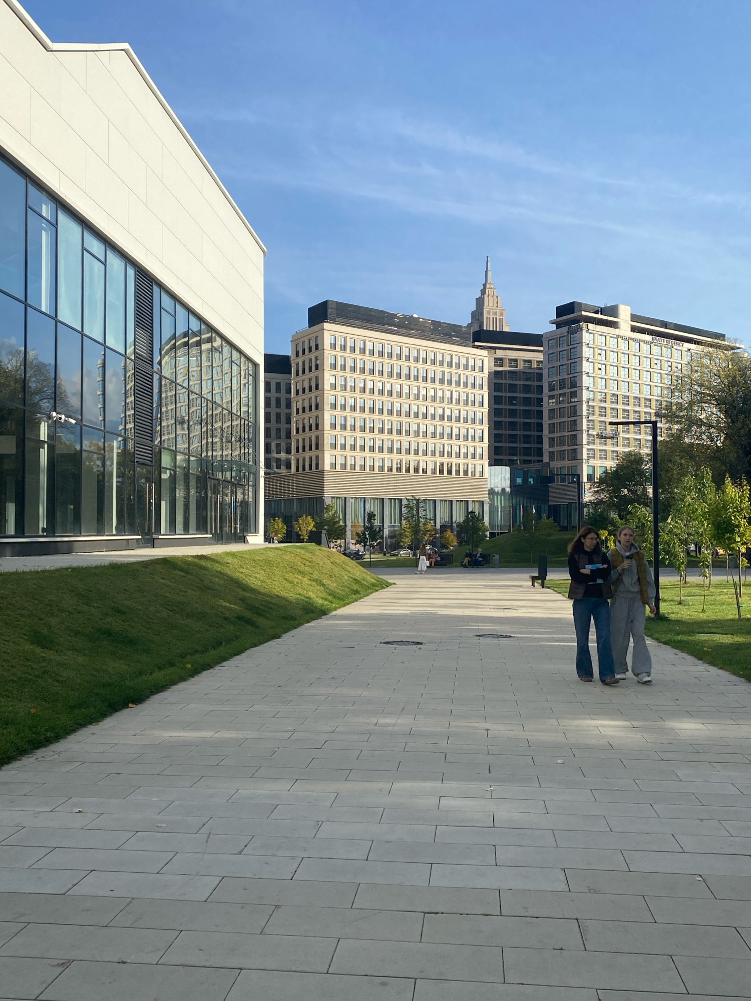 In a city park in Unknown, Unknown, two women walk on a paved path. The woman on the left wears blue jeans and a dark vest over a lighter shirt, while the woman on the right wears a gray tracksuit and a beige vest. They are walking towards the viewer. Behind them, a light-colored building with large glass windows reflects the sky and other buildings. Further in the background, several taller, modern buildings with many windows rise against a clear blue sky with wispy clouds. A prominent spire-like structure is visible atop one of the buildings. Green grass slopes gently up to the left of the paved path, and trees with green and yellow leaves are scattered throughout the park. The scene appears to be a bright, sunny day.