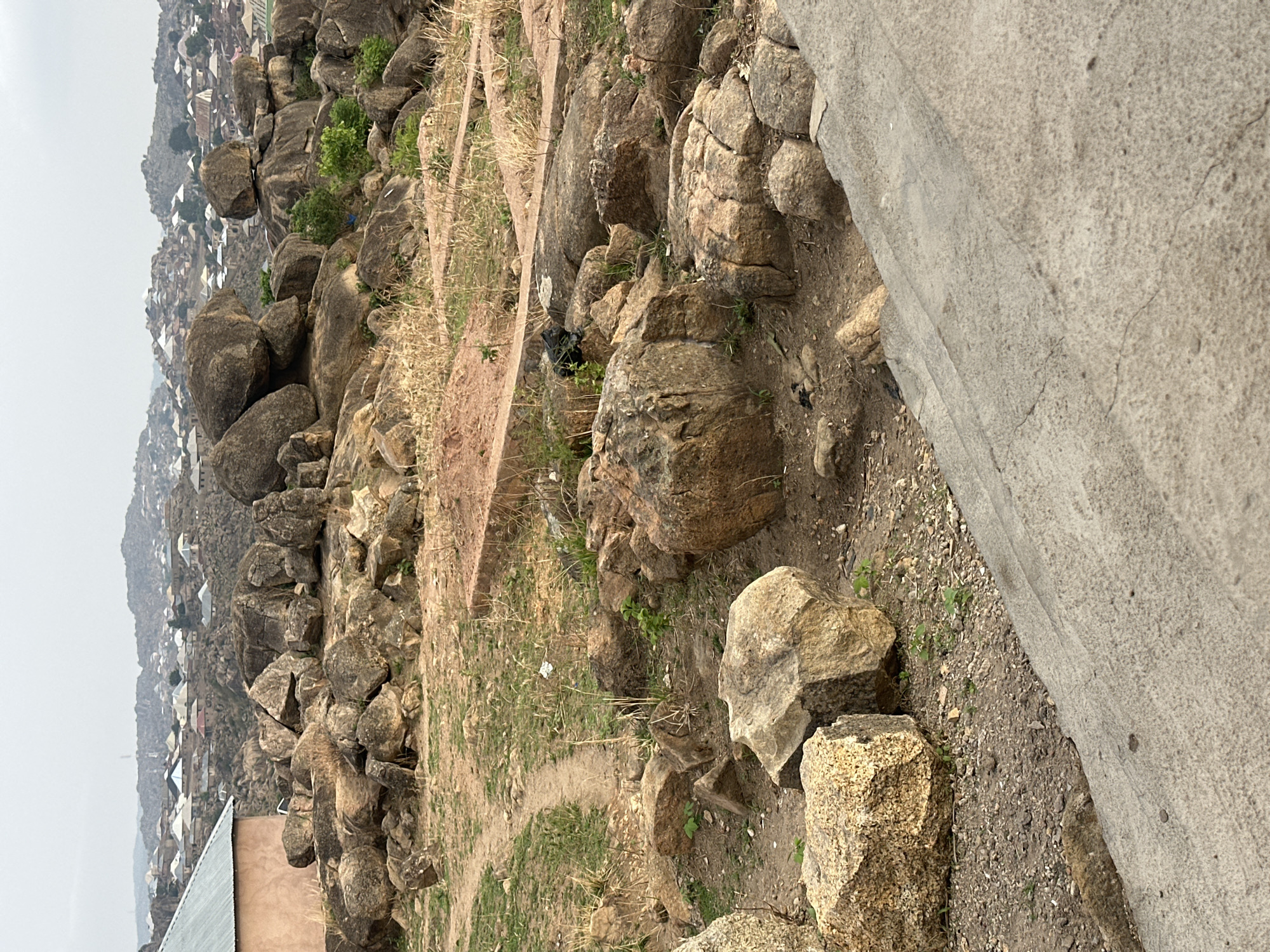 The image captures a rocky, elevated landscape with a dense urban settlement in the background, indicating a location near or within a mountainous region. Weather is hazy and overcast.
**Foreground:** A mix of rocks and boulders of varying sizes dominates the foreground. The rocks appear to be made of granite or a similar stone type, with textures ranging from smooth to rough. Patches of grass and scattered dry vegetation are visible between the rocks. A concrete structure of uncertain purpose or extension is visible on the right side, creating a boundary or edge.
**Background:** A densely built-up urban area stretches into the distance, with houses and buildings clustered together. There are hints of rooftops, walls, and other architectural details, which suggest a well-populated area. The terrain descends into the city, possibly a valley setting, viewed from a higher vantage point.
The general features indicate the vicinity of Jos, Nigeria, owing to the city's characteristic rock formations and mountainous landscape.