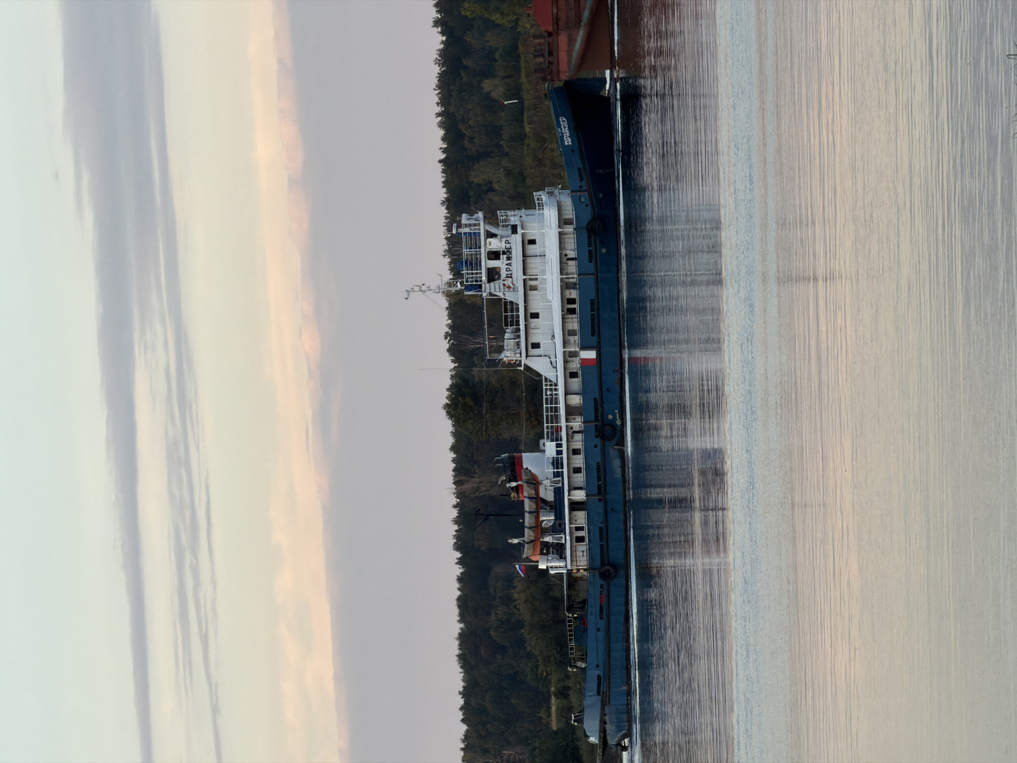A blue and white tugboat named "ДРАЙЗЕР" is sailing on a body of water, likely a river, with a forested background. The tugboat has a white superstructure with multiple windows and a red and white striped funnel. A Russian flag is visible on the deck. To the right of the tugboat, a portion of a red barge is visible, indicating the tugboat may be towing or pushing it. The water's surface reflects the sky and the boat, with ripples creating a textured effect. The sky above is a soft blend of pale blue and pink, with thin, wispy clouds. The overall scene suggests a calm, possibly evening, setting on a waterway near a wooded area. The city of Chernushka, Russia, is known for its rivers and proximity to forests, making this scene plausible for the region.