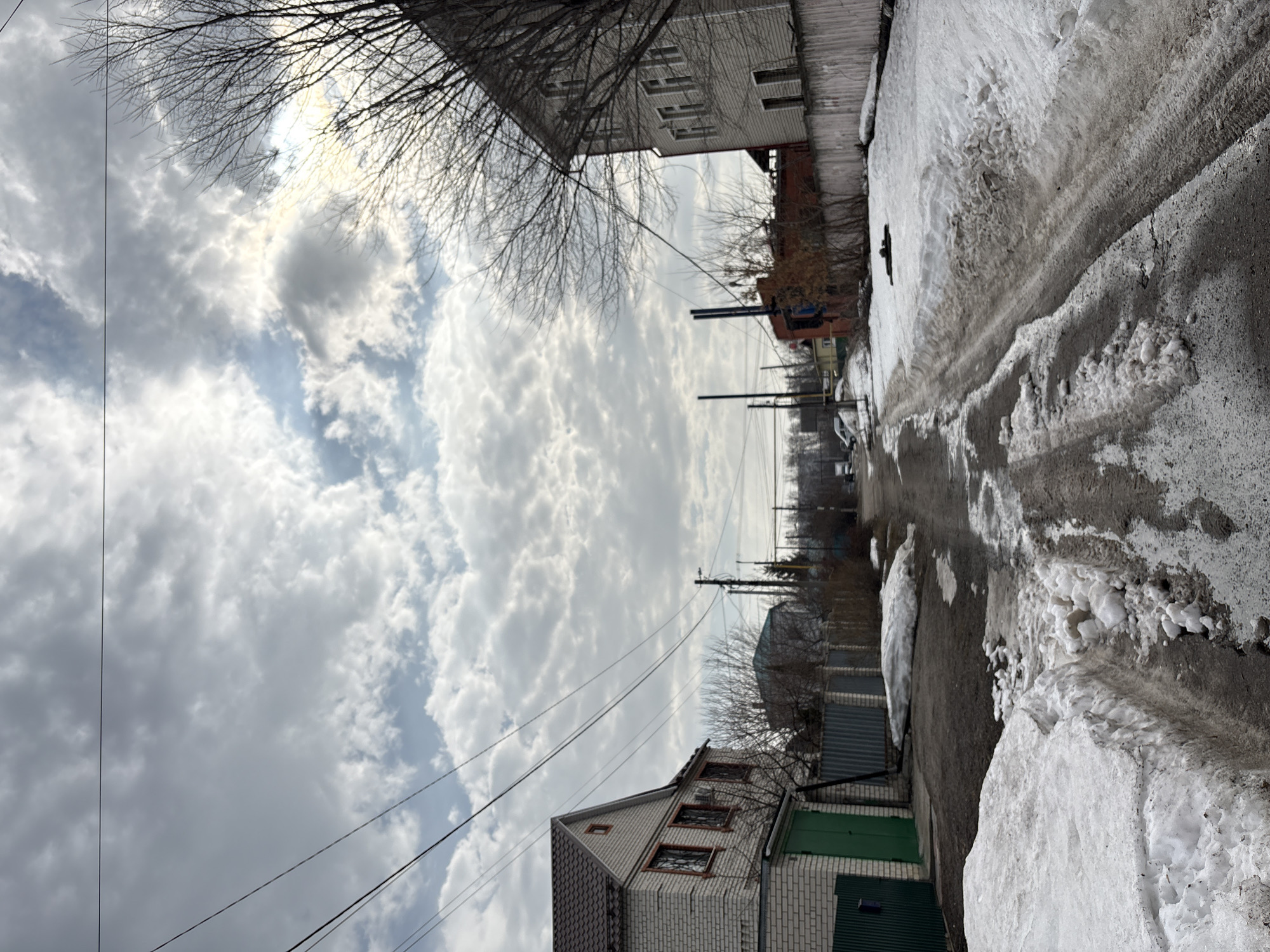 The image shows a street scene in Ulyanovsk, Russia, in what appears to be late winter or early spring. The street is narrow and paved, with melted snow and ice patches. Snow is piled up along the sides of the road. Two-story brick buildings border the street. Power lines run overhead. The sky is overcast with clouds.