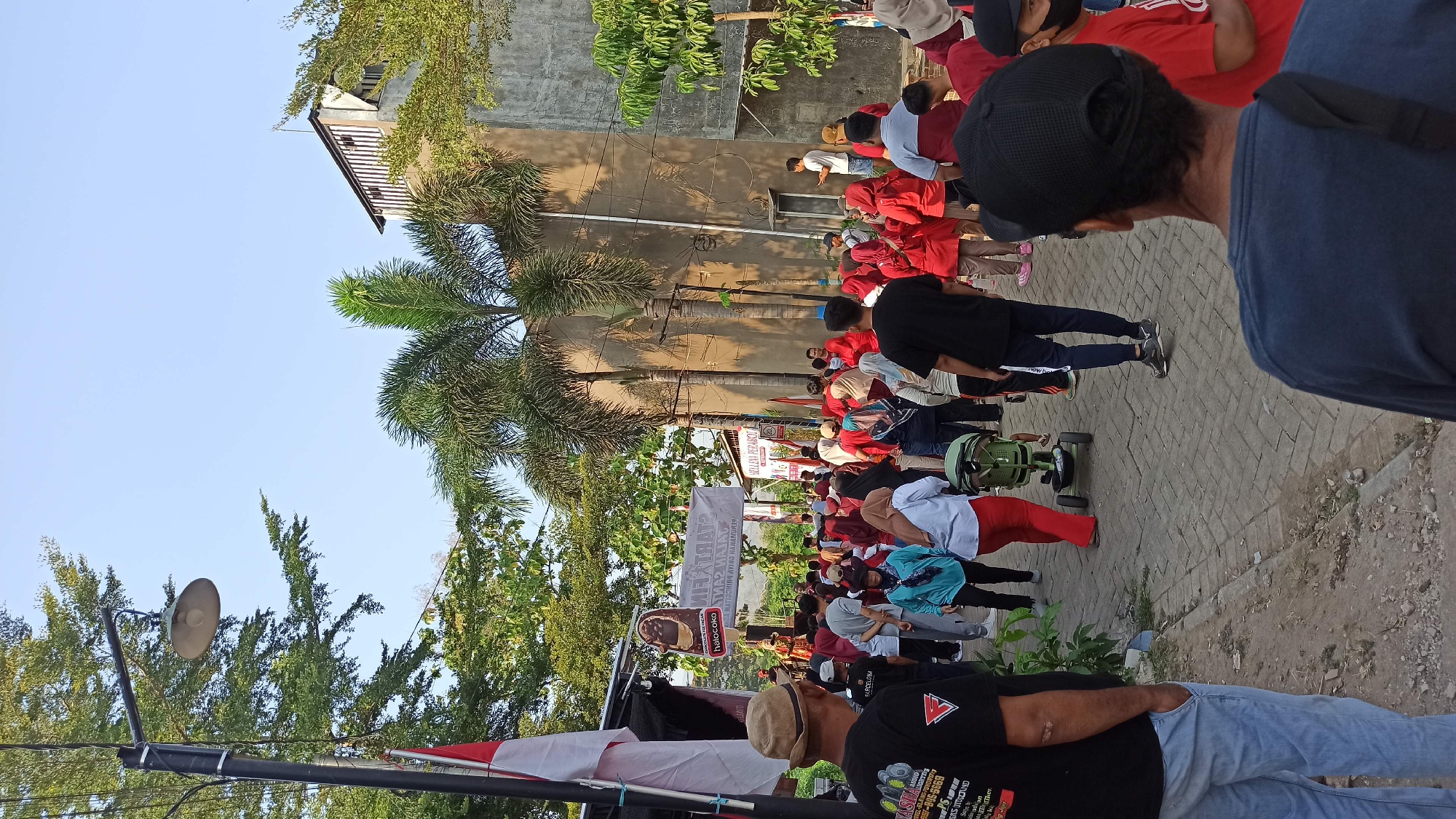 The image depicts an outdoor gathering of people in Galak, Indonesia, possibly for a community event or festival. A group of individuals, many wearing red clothing, are congregated on a brick-paved street lined with trees and buildings. A banner with Indonesian text, possibly advertising the event, is visible in the background, along with a flag. In the foreground, several people are looking towards the gathering, with one person wearing a black t-shirt and a straw hat in the left of the frame, and another wearing a blue t-shirt and a black baseball cap in the right. A small green stroller is also present in the middle of the scene. The overall atmosphere appears to be lively and communal.