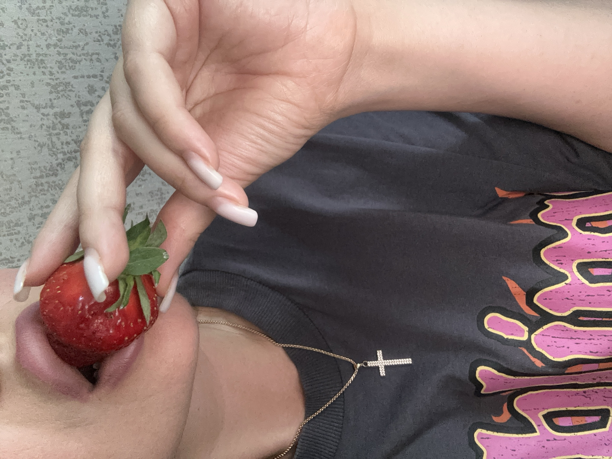 The image shows a person holding a strawberry to their mouth. The person has long, white fingernails and is wearing a gray t-shirt with a pink and orange design. A gold necklace with a cross pendant is visible around their neck. The background appears to be a textured wall. There is no indication that this image was taken in the city of Chaikovsky, Russia.