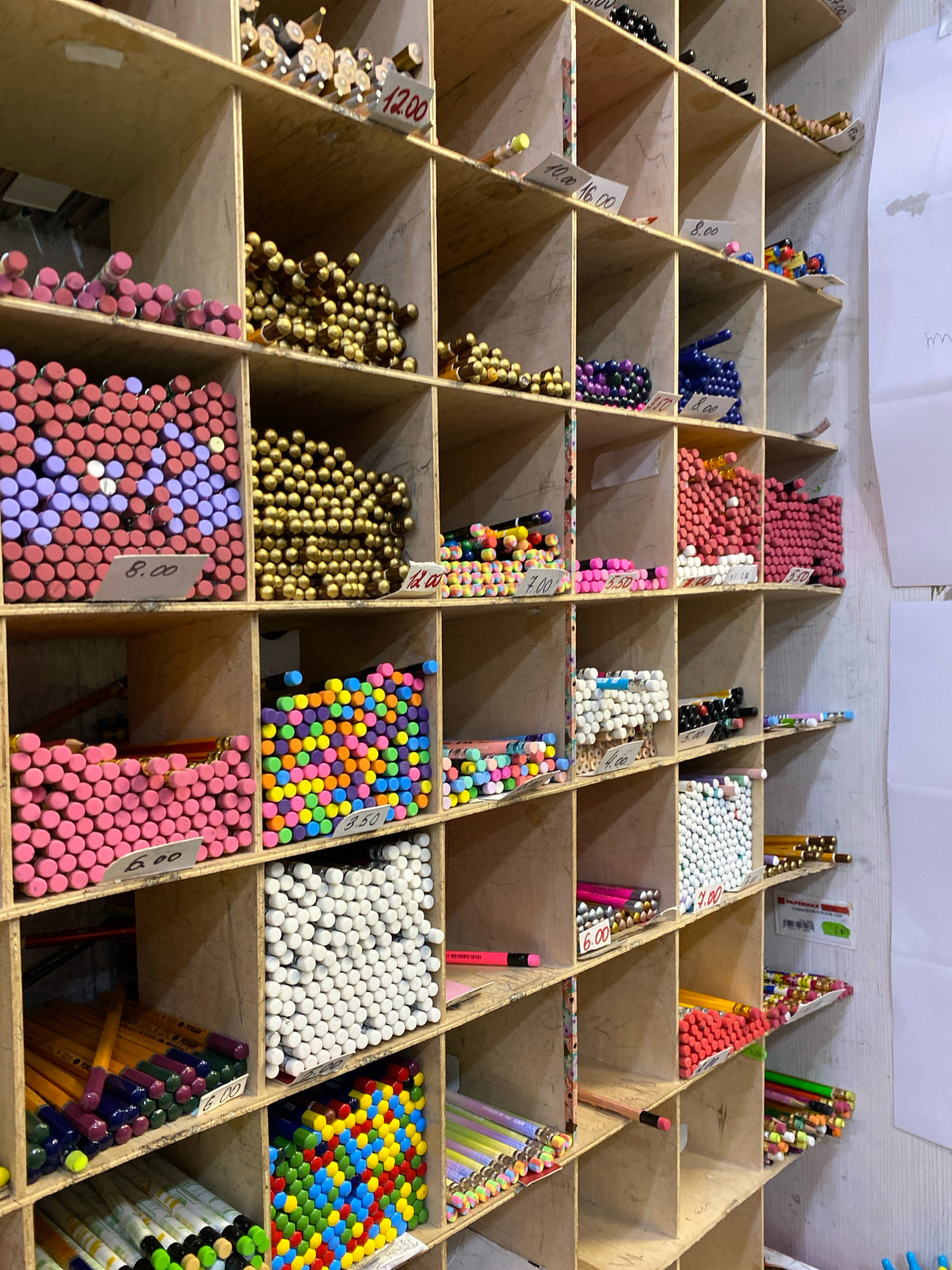 The image shows a retail display of art supplies, likely pencils, in a store in Chișinău, Moldova. The display is made of rough wooden shelves divided into numerous cubbies, each filled with colored pencils sorted by hue. Prices are marked on small white tags within some of the cubbies. The lighting is artificial and casts shadows. The shelves are packed with a variety of colored pencils in bulk, neatly arranged. The overall impression is of a well-stocked art supply store.