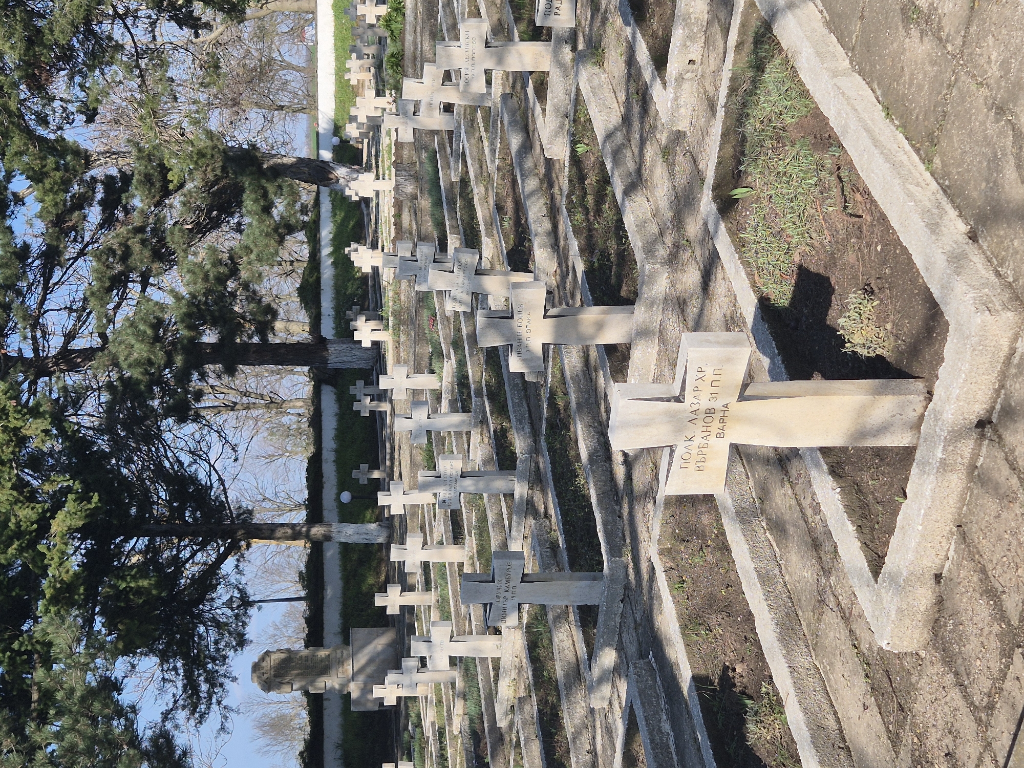 The image shows a cemetery with rows of similar-looking, light-colored stone grave markers. The markers are mostly in the shape of crosses. Trees are visible near the left side of the image, and the sky is visible in the upper left. The markers are surrounded by concrete frames with barren or partly vegetated ground in between. Text is inscribed on many of the markers, appearing to be in Bulgarian. One marker reads "ПОЛК. ЛАЗАР ХР. ВЪРБАНОВ 31 П.П. BAPHA". The image is rotated. The location may be Shumentsi, Bulgaria.