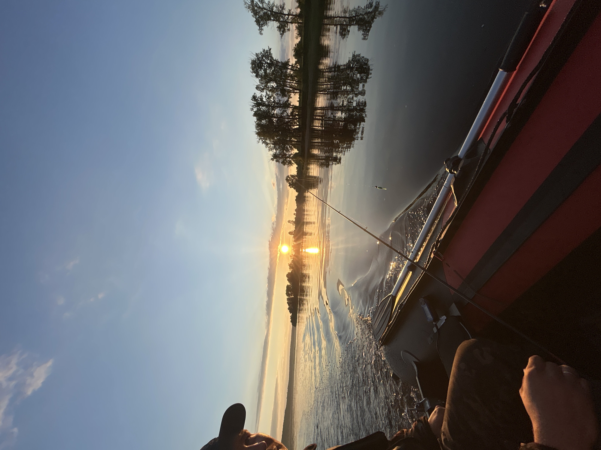 An eye-level, medium shot captures a person fishing from an inflatable boat on a calm body of water at sunset. The person, wearing a dark cap and jacket, is seated on the left side of the boat, facing away from the camera. Their hands are visible, one resting on their lap and the other near a fishing rod. The boat itself is red and black and contains oars. A fishing rod extends over the side of the boat, with a bobber visible in the water. The sun is setting on the horizon, casting a warm glow across the water and illuminating the sky with soft orange and yellow hues. Distant trees and a shoreline are visible across the water, their reflections shimmering on the water's surface. The overall scene conveys a sense of peace and tranquility. The location is identified as Ol'khovskoye, Russia.