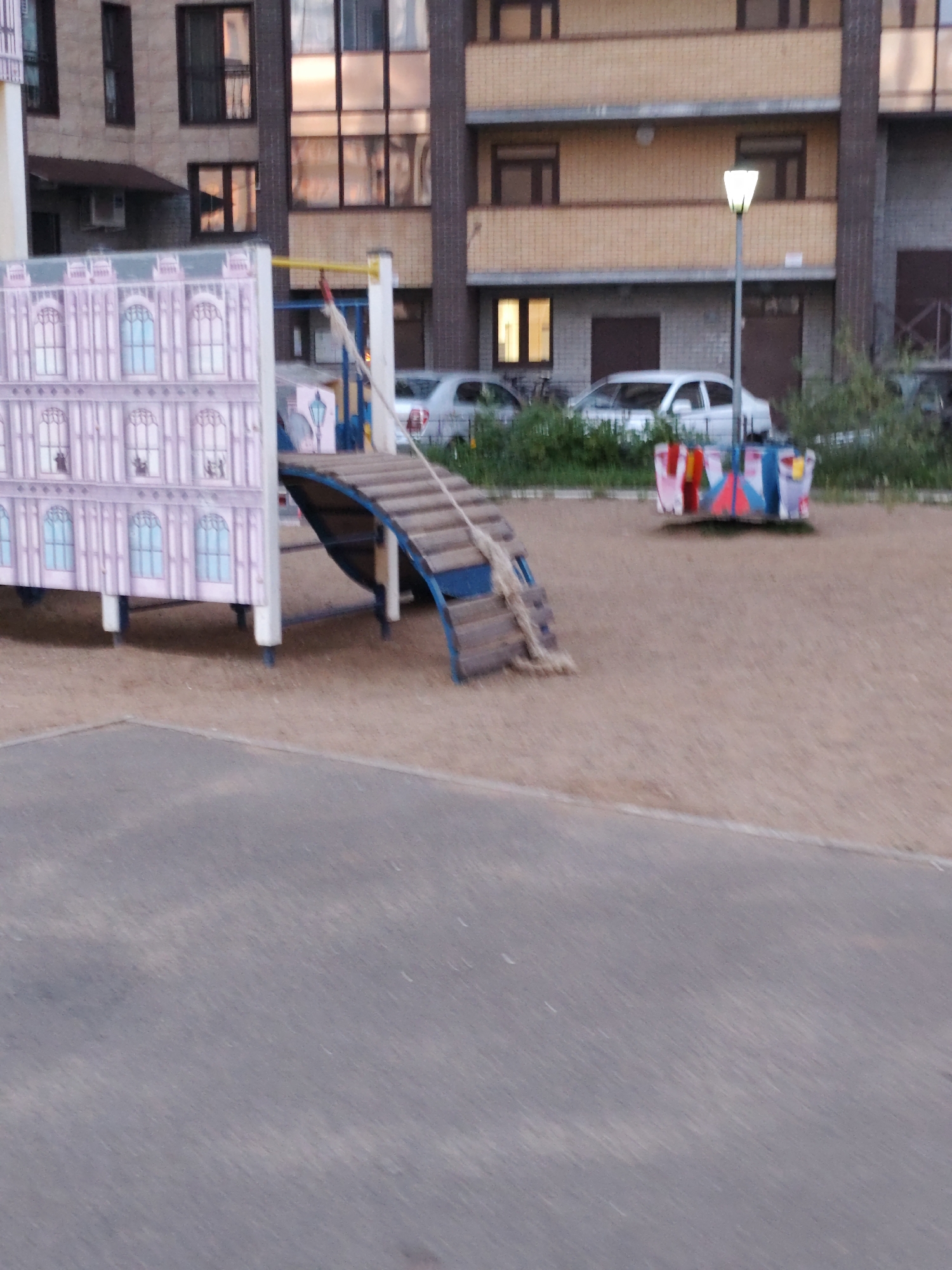 The image depicts a courtyard of a residential building in Syktyvkar, Russia. In the foreground, there is a paved area. Behind it, a playground with a slide and a merry-go-round is visible. The slide is made of wood and rope, and the merry-go-round has colorful seats. The residential building is a multi-story structure with a brick facade and large windows. The building appears to be modern, with balconies on some floors. Several cars are parked in front of the building, and there is a street lamp in the background. The overall scene suggests a typical urban environment, likely in the late afternoon or early evening, given the lighting conditions. The playground is the central focus of the image, implying it is a communal space for children within the residential complex.