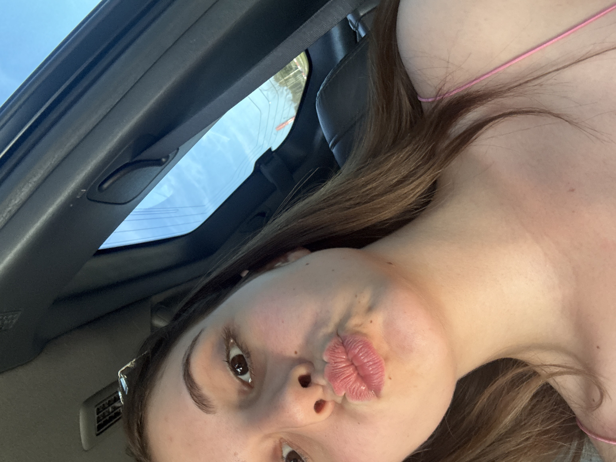 A young Caucasian female is taking a selfie from inside a vehicle, likely a car. She is looking directly at the camera with her lips pursed in a "duck face" pose. Her eyes are open, and she has brown hair. She is wearing a thin pink strap, suggesting she is wearing a camisole or tank top. The background shows the interior of the car, including the ceiling, a window with a view of the sky and some distant trees, and car seats. The lighting suggests it is daytime. There is no information in the image to suggest a specific location in Burgas, Bulgaria.