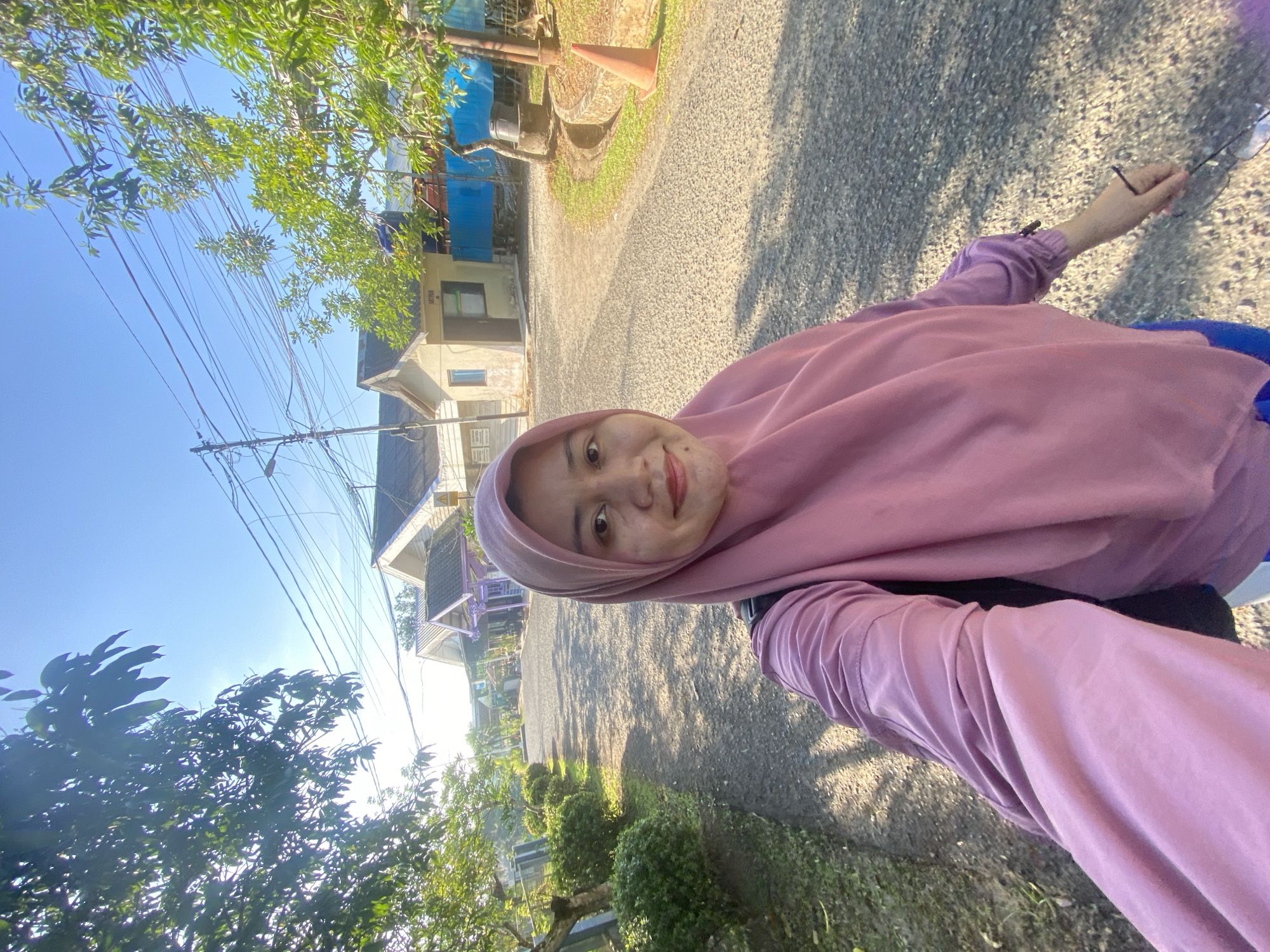 A woman wearing a pink hijab and a pink top is standing on a gravel path. She is holding a pair of glasses and a purple object in her right hand. The woman is looking at the camera with a smile. In the background, there are houses, trees, and power lines. The scene appears to be in a rural or suburban area. The image is taken outdoors during the day. The location is specified as Banjarbaru, Indonesia.