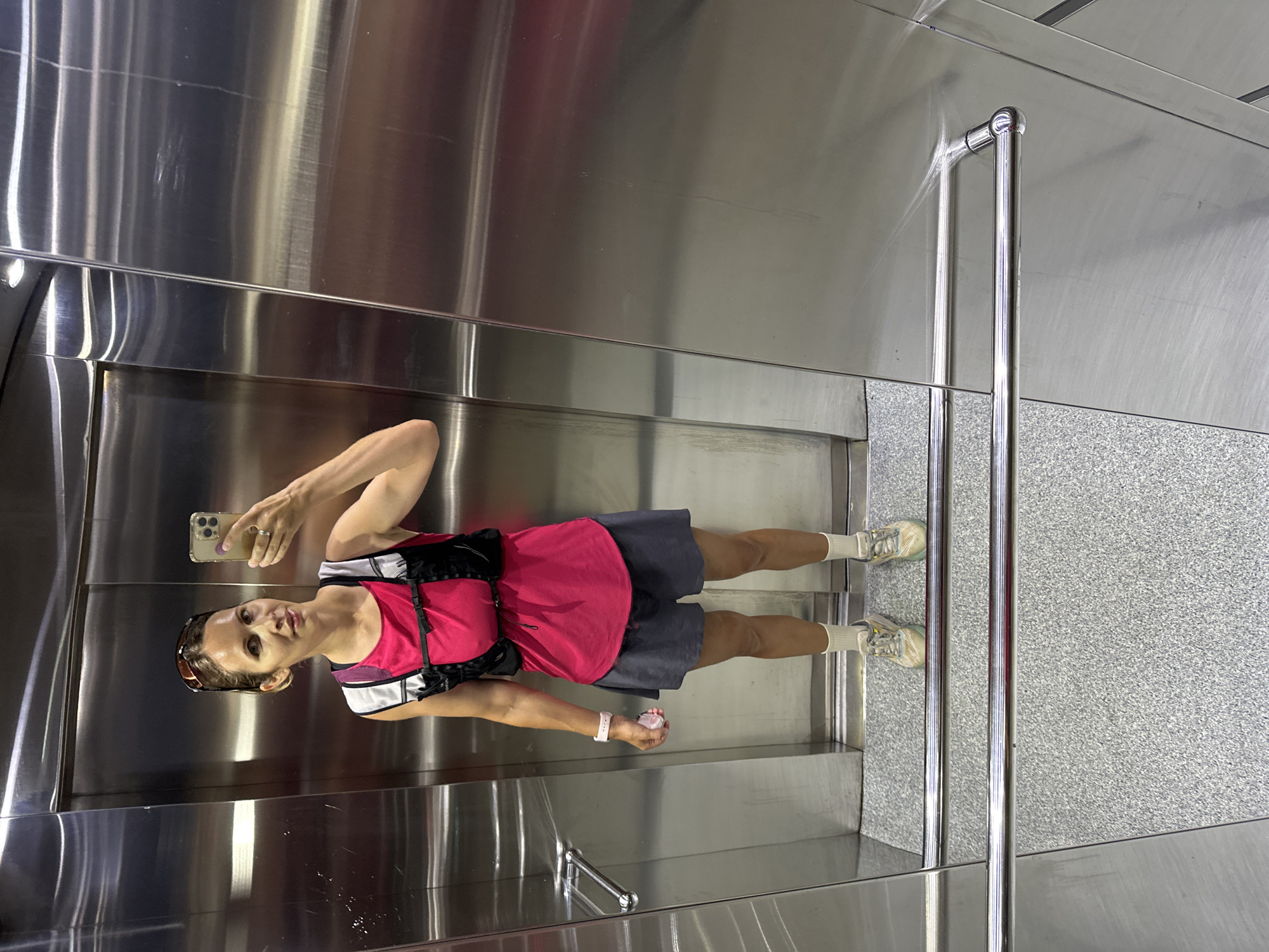 A woman is standing in what appears to be an elevator. She is facing the camera, holding a cell phone in her left hand, and seems to be taking a selfie. She is wearing a pink top, dark shorts, white socks, and sneakers. The elevator interior is primarily composed of stainless steel with a textured floor. There is a vertical stainless steel grab bar near the front of the elevator.
It is impossible to determine whether this image was taken in the city of Fethiye, Turkey.