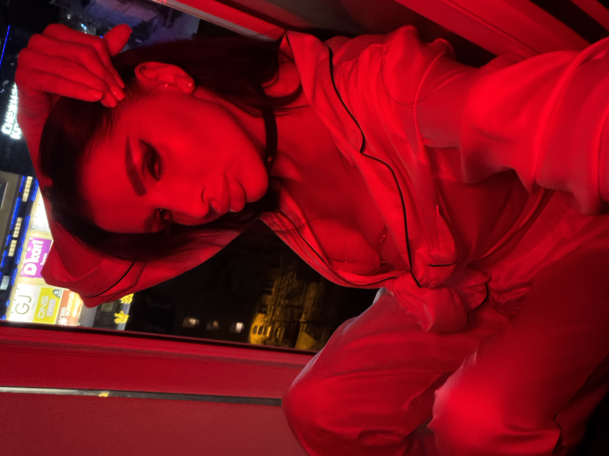 A person is indoors, near a window, bathed in red light. The person is wearing what appears to be a satin pajama set with dark trim. They are touching their hair with one hand. Outside the window are buildings, including one with a sign that reads "DKart" and "DNS", indicating a commercial area. The city is Svkh Voronezhskiy, Russia.