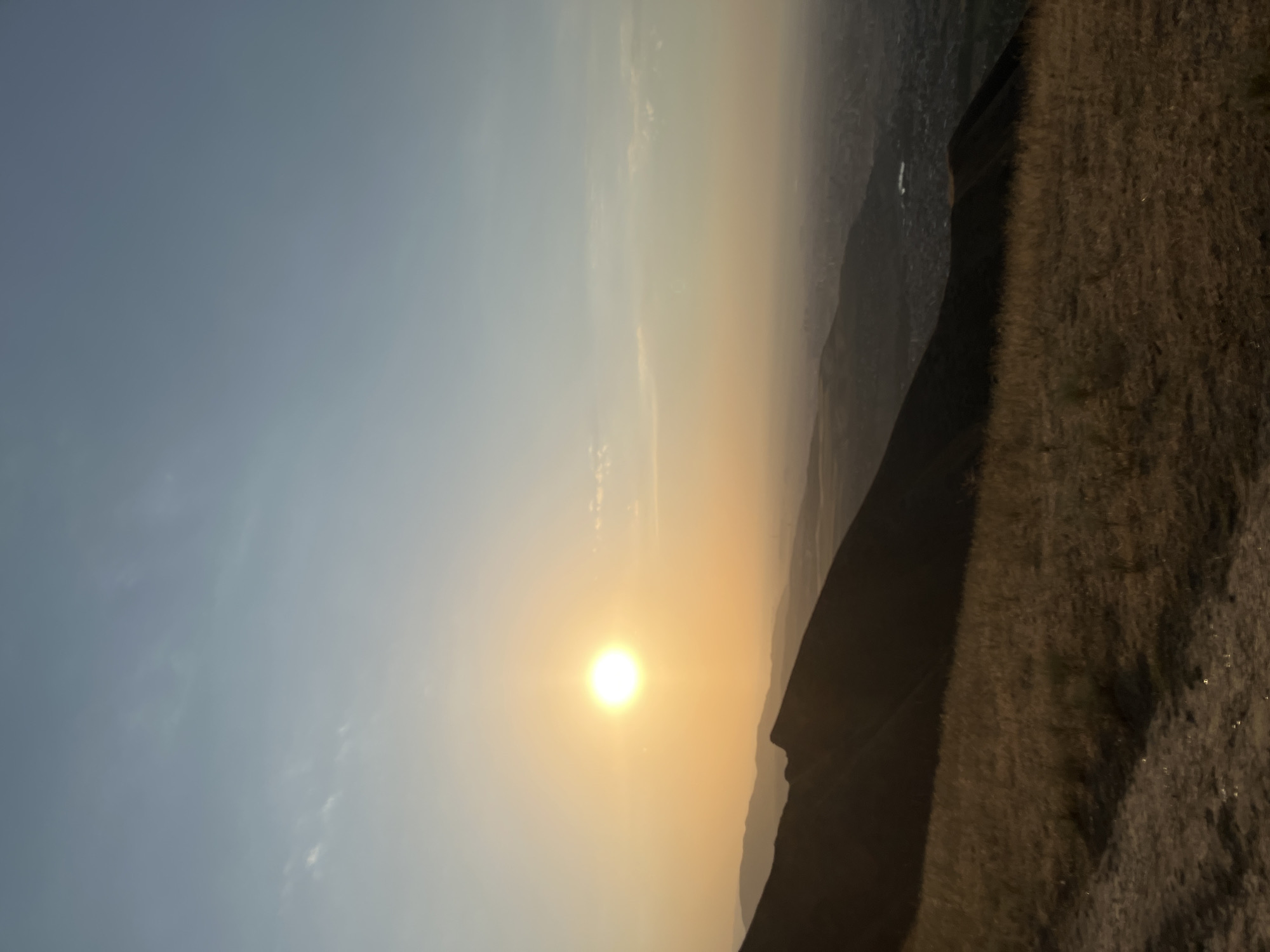 The image depicts a panoramic view from a hilltop overlooking Bishkek City, Kyrgyzstan, at sunset. The sun, a bright, glowing orb, hangs low in the sky, casting a warm, golden-orange hue across the landscape. Wisps of thin clouds are scattered across the clear, pale blue sky. In the foreground, a grassy slope covered in dry, yellowed grass leads down towards a dark, silhouetted mountain ridge that cuts across the frame. The ridge has a rugged, sharp outline. Beyond the ridge, the city of Bishkek sprawls across the valley, its buildings and lights faintly visible through a hazy atmospheric perspective. The overall impression is one of serene natural beauty combined with the distant presence of urban life.