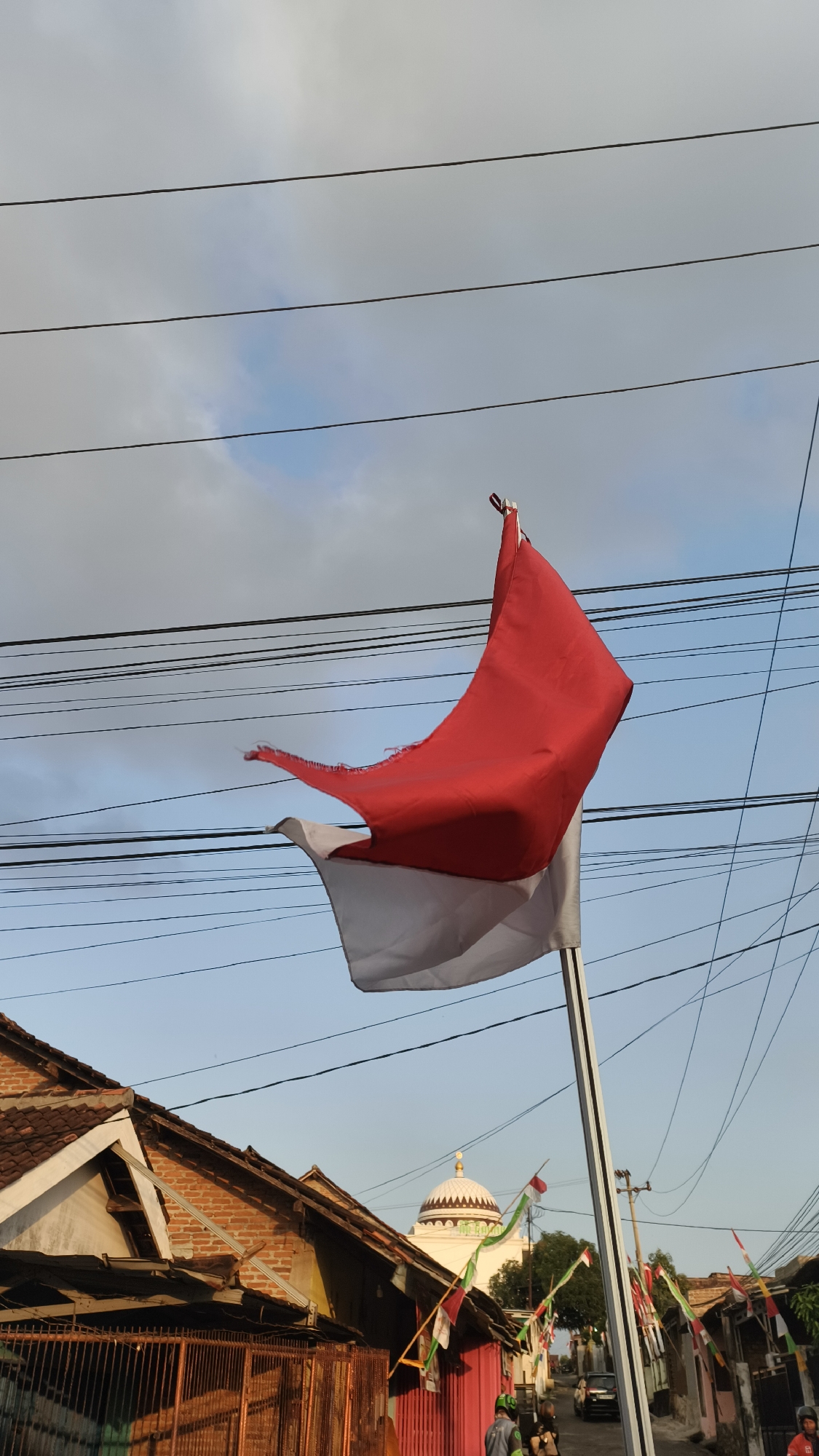 An Indonesian flag, red and white, waves on a pole against a cloudy blue sky. Several power lines crisscross the sky. In the background, a street is lined with houses, some with tiled roofs and brick walls. A building with a white dome, possibly a mosque, is visible in the distance. Small Indonesian flags adorn some of the houses and poles along the street. The scene appears to be a residential area in Bandar Lampung, Indonesia.