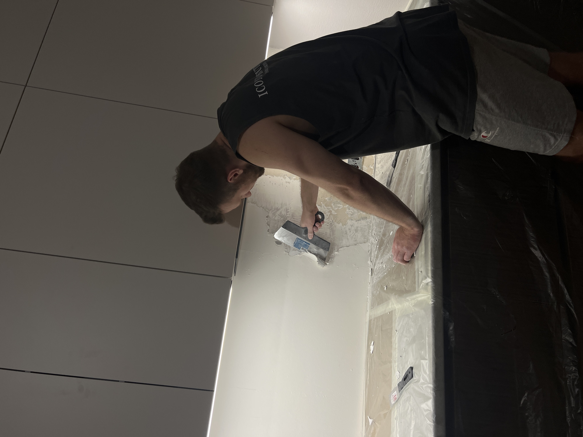 A man is using a trowel to apply plaster to a wall. He is wearing a black tank top and gray shorts. The wall is white and appears to be partially plastered. The floor is covered in plastic sheeting. The location in the city of Svkh Voronezhskiy, Russia is not identifiable from the image.