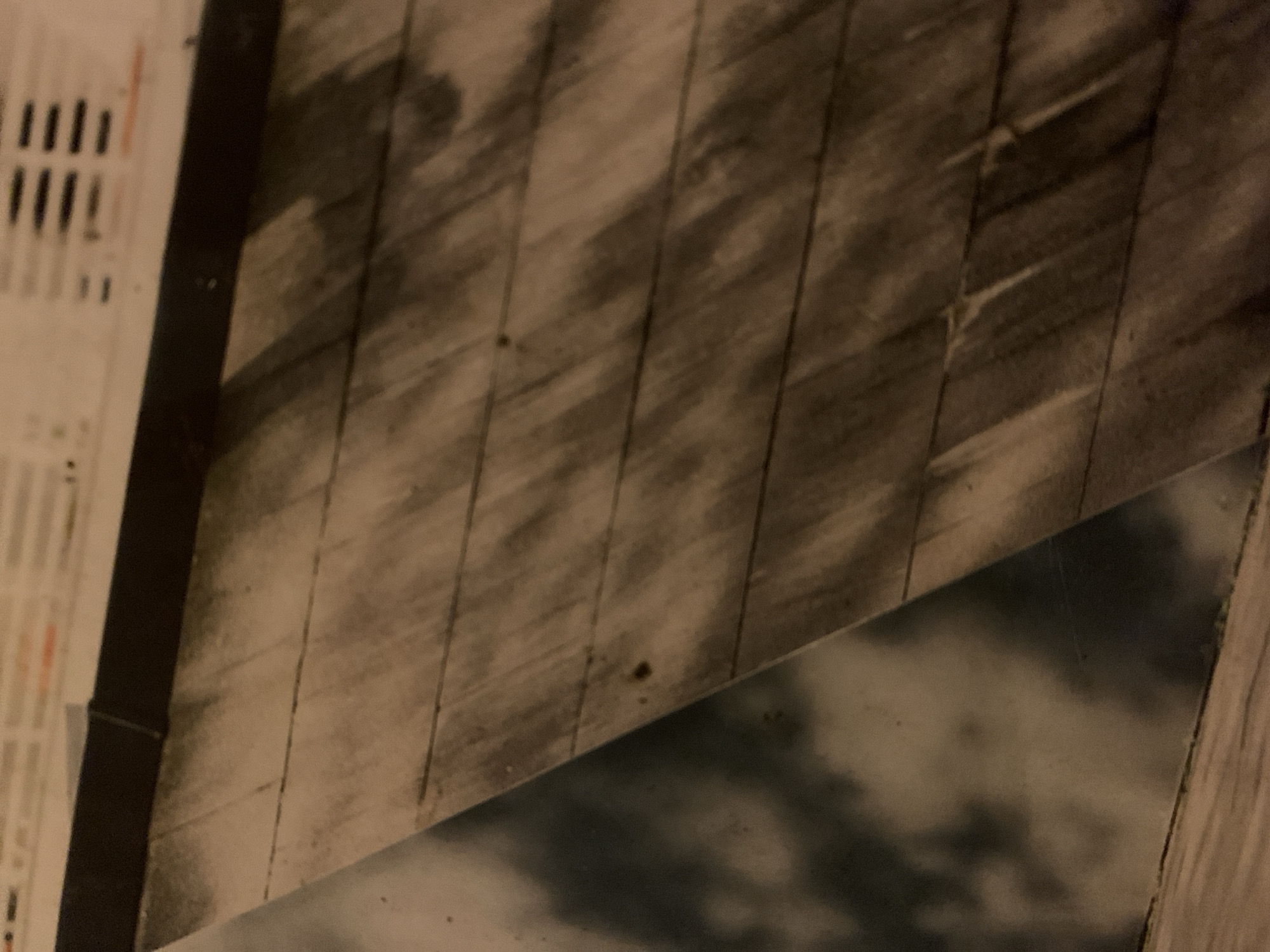 The image shows a close-up of a wooden wall or structure, with a rectangular piece of what appears to be a photograph or print of clouds partially visible. The wooden structure consists of narrow, vertical planks. To the left is a strip of what may be printed text or an image with lines and shapes. The photograph does not appear to be taken in Barcelona, Spain.