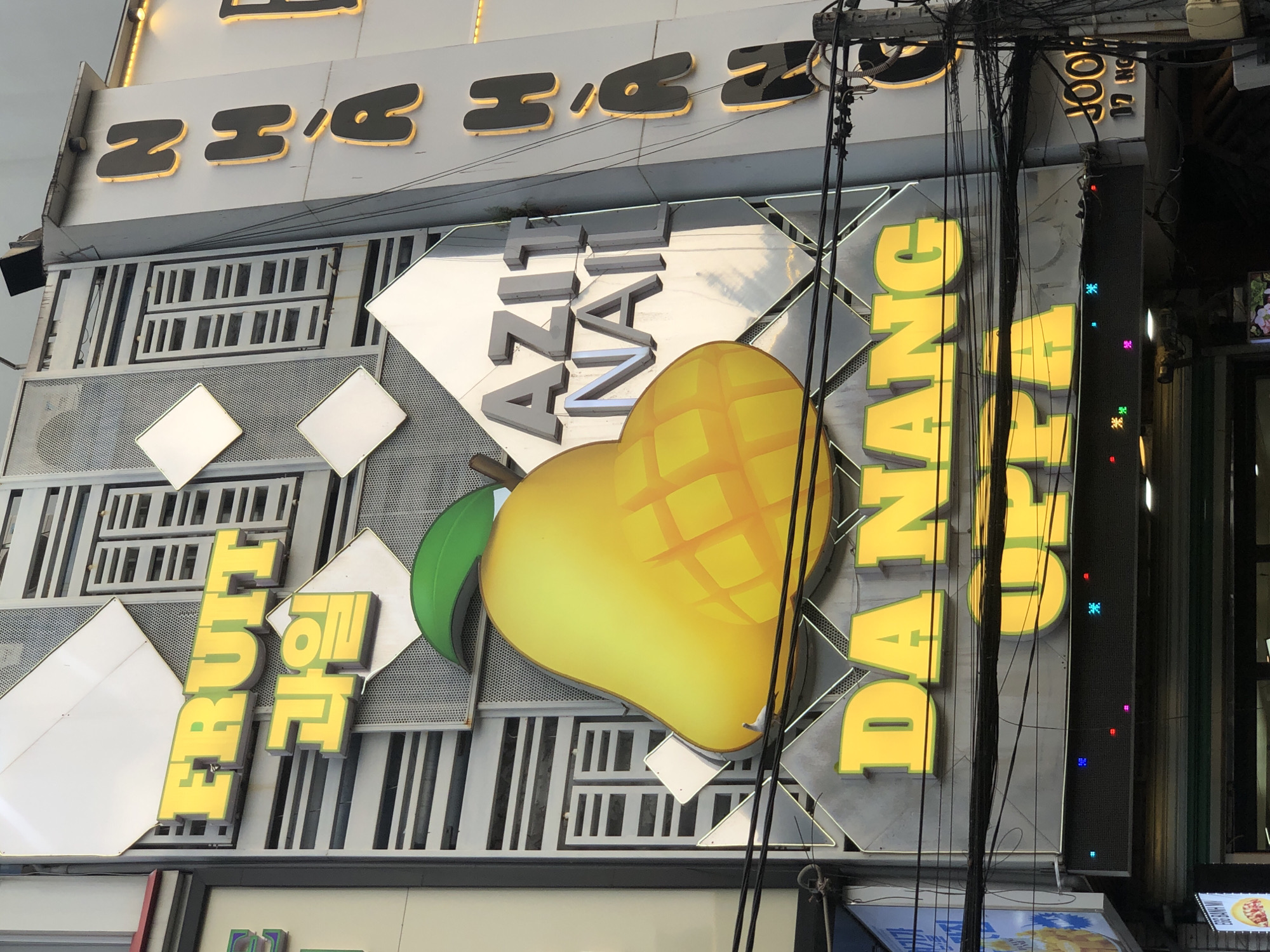 An exterior shot captures a brightly lit sign for "FRUIT" in yellow 3D letters with a Korean inscription below it. To the right, a large, glossy yellow mango illustration dominates the frame, with a green leaf sprout. Adjacent to the mango, silver 3D letters spell out "AZIT NAIL." Further to the right, a vertical sign displays dark, backlit letters: "N H À H À N." Below the mango and "AZIT NAIL," a prominent yellow sign reads "DA NANG." Beneath that, another yellow sign, partially obscured by wires, reads "UFPA." A horizontal LED display flashes with small colored lights. The background consists of a modern building facade with metallic and striped elements. A complex network of electrical wires crisscrosses the foreground. The image appears to be taken in a commercial district of Đà Nẵng, Vietnam, given the signage.