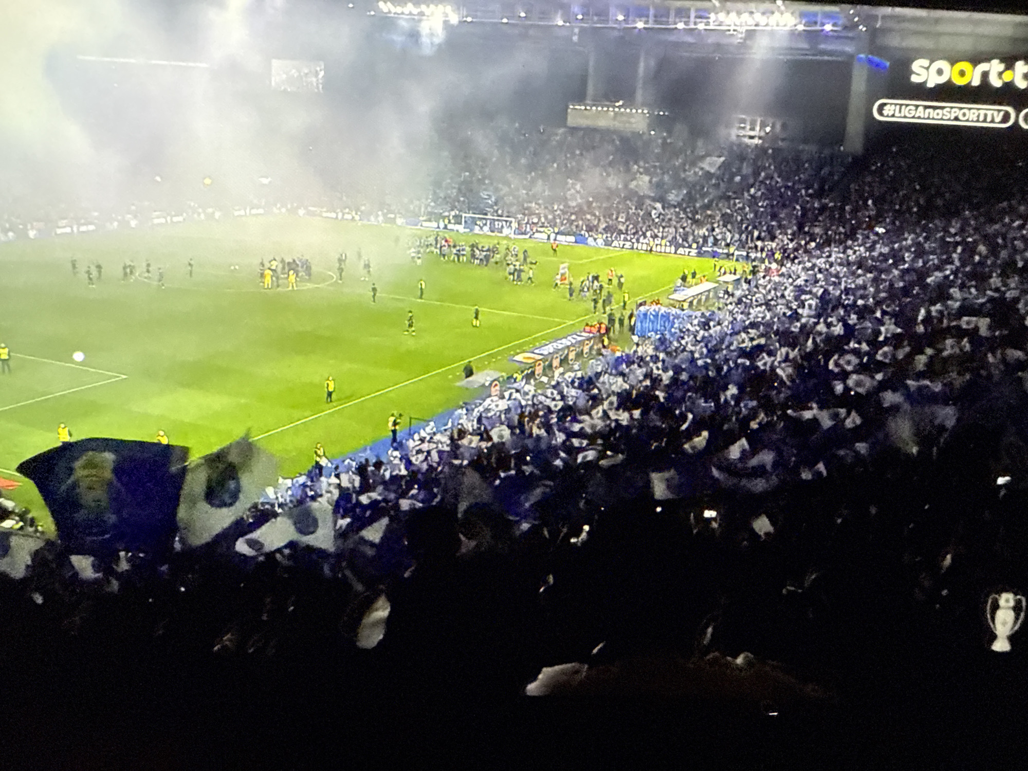 The image depicts a soccer match at the Estádio do Dragão, the home stadium of FC Porto in Vila Nova de Gaia, Portugal. The stadium is filled with fans, many waving white and blue flags. Smoke fills the air, obscuring parts of the field and stands. Players can be seen on the green pitch. The stadium's electronic signage is visible in the upper right corner, displaying "sport-tv" and "#LIGAnoSPORTTV." A Champions League trophy replica is in the bottom right corner. The scene suggests a celebratory atmosphere.