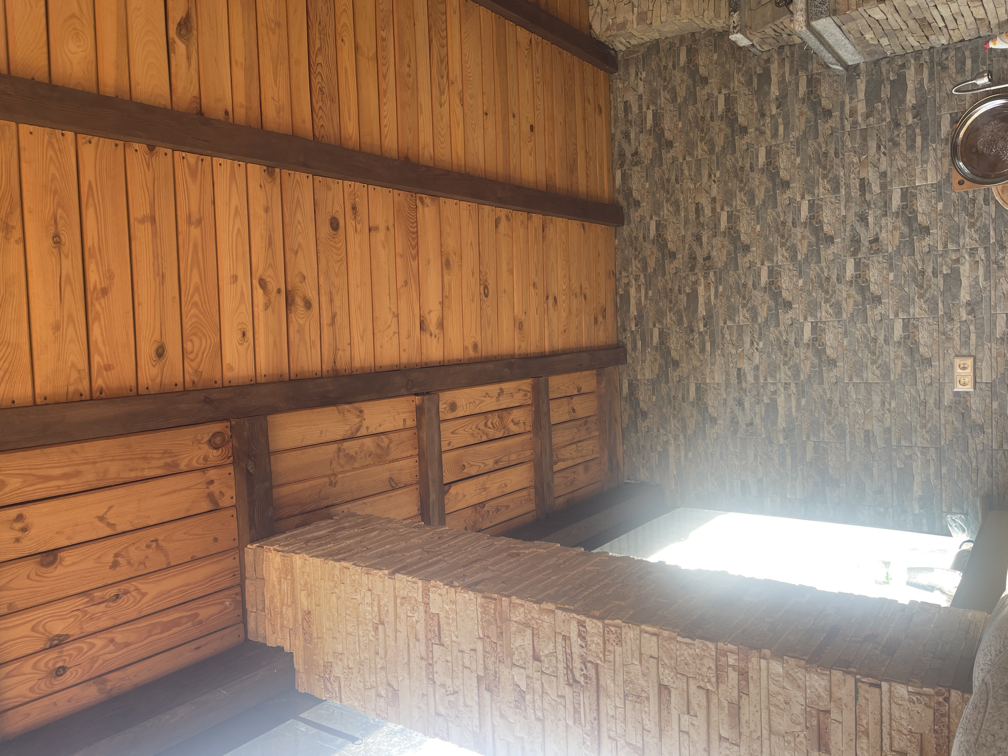 This image, located in Petrivka, Ukraine, captures a view of a covered outdoor or interior space, primarily focusing on its architectural elements from a low-angle perspective looking upwards. The dominant feature is the ceiling, constructed from numerous light-to-medium brown wooden planks laid horizontally, secured by visible nails. These planks are supported by several thicker, dark brown exposed wooden beams that run perpendicularly across the ceiling.
On the left side of the frame, a large, vertical structural pillar is visible, constructed from light-toned, stacked stone veneer with a rough, irregular texture. This pillar is heavily backlit by intense, blinding sunlight streaming from an unseen opening or window on the far left, creating a strong glare and obscuring details beyond.
The wall on the right and across the background is also clad in stacked stone veneer, featuring a mixture of gray and brown irregular-shaped stones. Towards the lower left of this wall, two white dual electrical outlets are affixed. In the bottom right corner, a glimpse of a reflective, circular silver tray with embossed detailing is visible, placed on what appears to be a counter or surface, adjacent to a metal faucet or tap, suggesting a utility or kitchen area. No people are present in the scene. The overall environment conveys a rustic or natural architectural style under bright daylight.