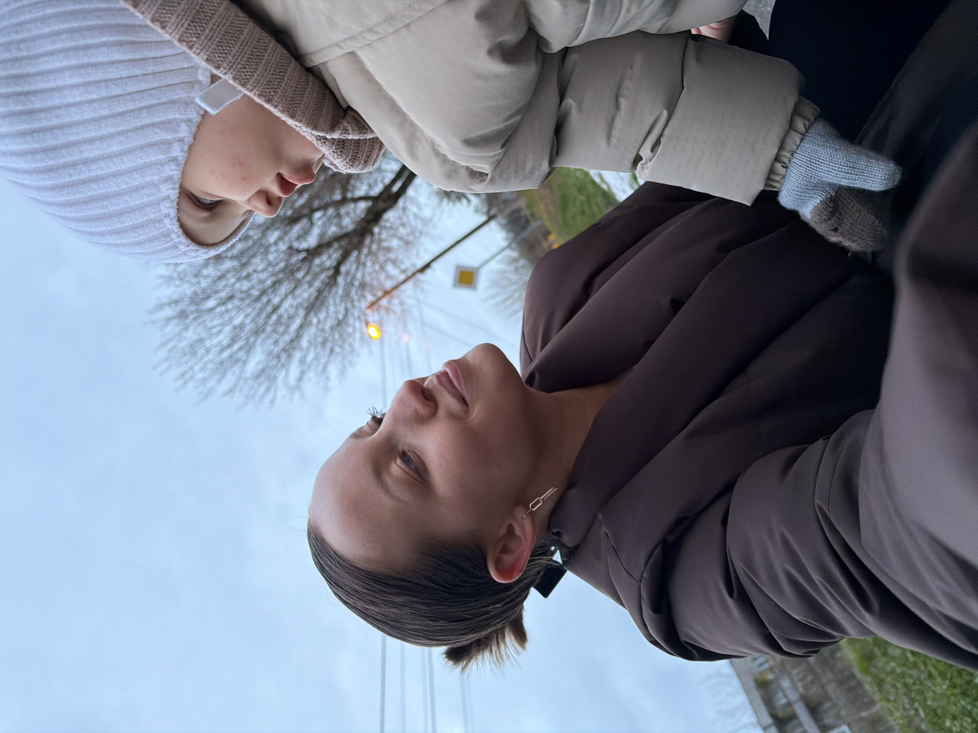 This image features two individuals outdoors, likely in Reshetnikovo, Russia, during cool or cold weather. The photo is taken from a low angle, looking upwards.
On the right side of the frame, an adult woman is visible from the chest up. She has dark hair pulled back and wears a dark brown puffy jacket and a gold-toned chain-link earring. Her expression is neutral as she looks generally forward, slightly to her left.
To her left, a younger person, likely a child, is visible from the chest up. They wear a light beige ribbed knitted hat and a light beige puffy jacket. Light blue knitted gloves with brown fingertips are also visible on their left hand. The child has light-colored eyes and is looking slightly upwards and towards the woman.
The background consists primarily of a dull, overcast sky, suggesting an overcast day. Bare tree branches are visible against the sky, indicating a late autumn or winter season. In the distance, a yellow diamond-shaped road sign, indicating a priority road, and a street light with a warm glow can be seen. Faint glimpses of distant buildings and patches of grass are also present. The individuals' attire (puffy jackets, knitted hat, gloves) strongly suggests chilly temperatures. There is no visible text in the image.