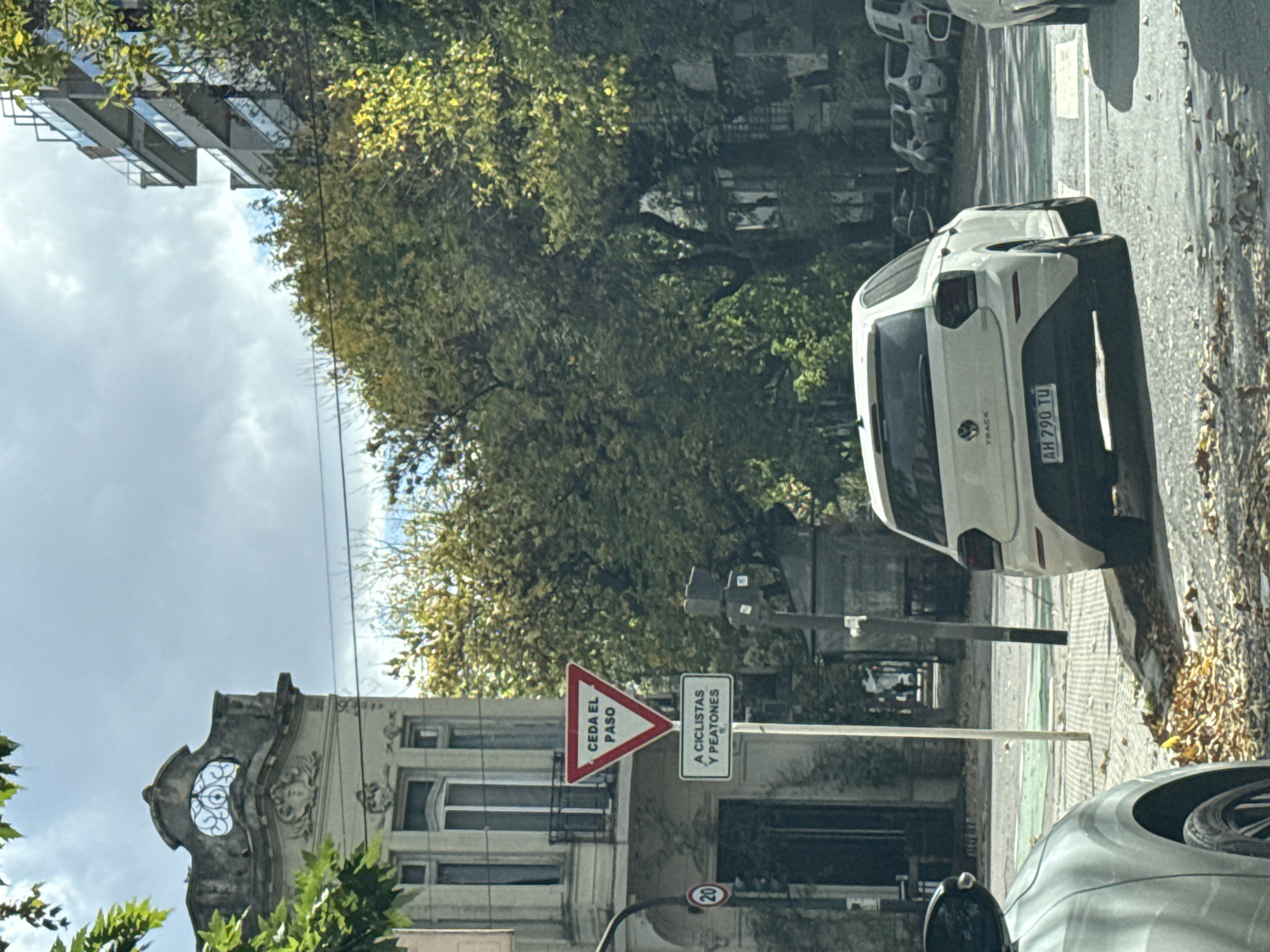 The image shows a street scene in Buenos Aires, Argentina. A white car is parked on the side of the road, a "Ceda El Paso" (Yield) sign indicating a crosswalk for pedestrians and cyclists is visible. The license plate of the white car reads "AH 790 TU". A building with ornate architectural details and an oval window is in the background, along with trees and other parked cars. The leaves on the street indicate that it might be autumn.