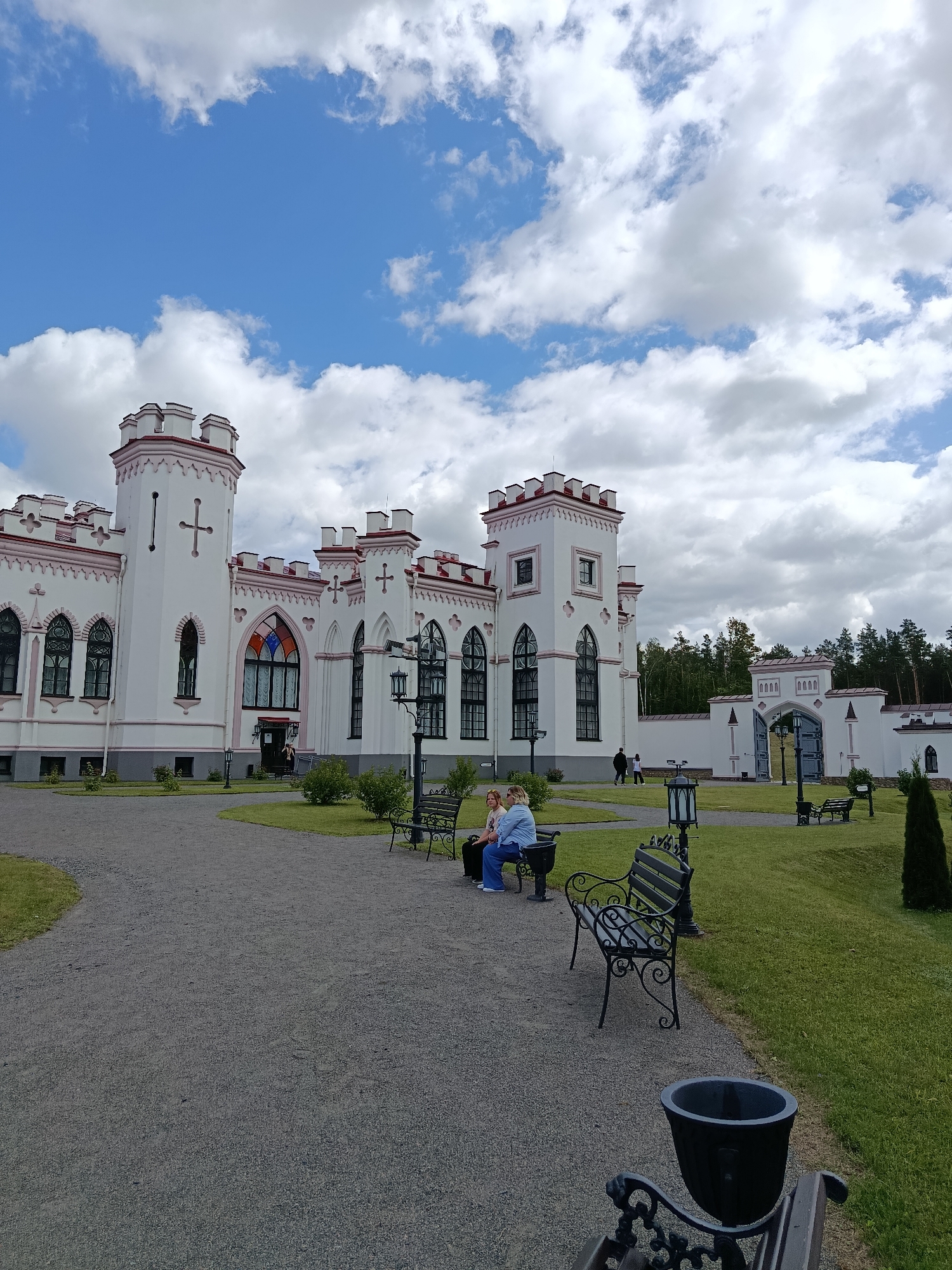 The image depicts the Kosava Castle in Kosava, Belarus, on a cloudy but bright day. The castle is a white, Gothic-style structure with crenellated towers and pink trim. A large gravel path leads to the castle entrance, which features a colorful stained-glass window. Several benches are placed along the path, and two women are seated on one of them, facing away from the camera. A large gate with an arched opening is visible in the background, along with a dense line of trees. The grounds are covered in grass and small bushes, and a dark trash receptacle is in the foreground.