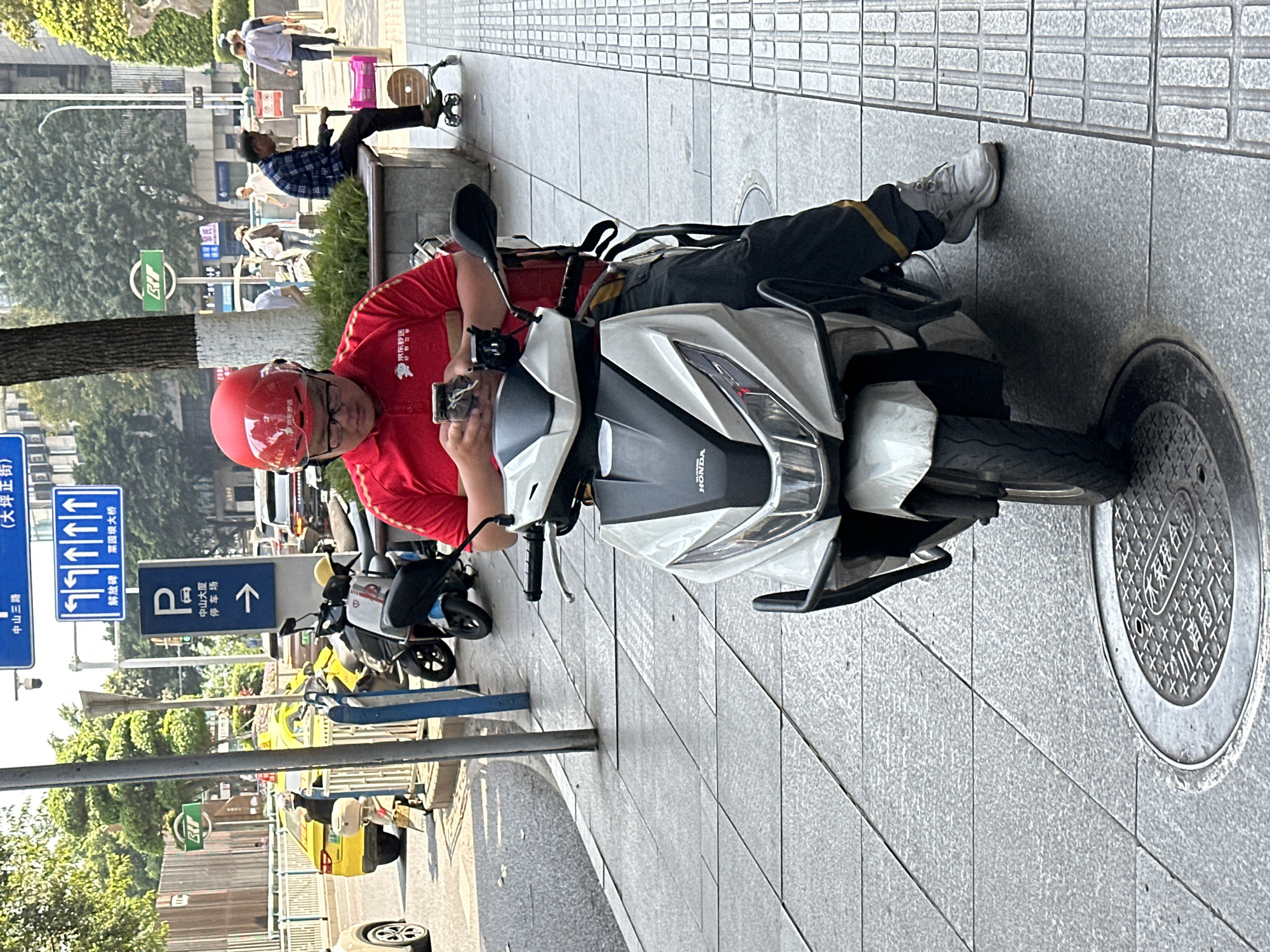 A delivery rider, dressed in a red short-sleeved uniform shirt with "京东秒送" (Jingdong Miaosong) printed on it, and a matching red helmet, is paused on a white and black Honda scooter. He is looking down at his smartphone, which he holds with both hands. He wears dark pants with a yellow stripe and light-colored sneakers.
The scene is an urban street or sidewalk in Yuzhong District, China, likely during the daytime under sunny conditions, indicated by clear lighting and shadows. In the background, other pedestrians walk along the sidewalk, and one person sits on a bench. Various vehicles, including cars and other scooters, are visible on the street.
Several street signs are present:
* A blue sign with multiple arrows indicates directions to "中山三路" (Zhongshan 3rd Road), "(大坪正街)" (Daping Zheng Street), "解放碑" (Jiefangbei), and "菜园坝大桥" (Caiyuanba Bridge).
* Another blue sign with a "P" symbol and an upward arrow points to a "停车场" (parking lot) for "中山大厦" (Zhongshan Building).
* A green sign with the letter "B" is partially visible.
* A manhole cover on the sidewalk displays Chinese characters, including "木结构台" and "川路管厂".